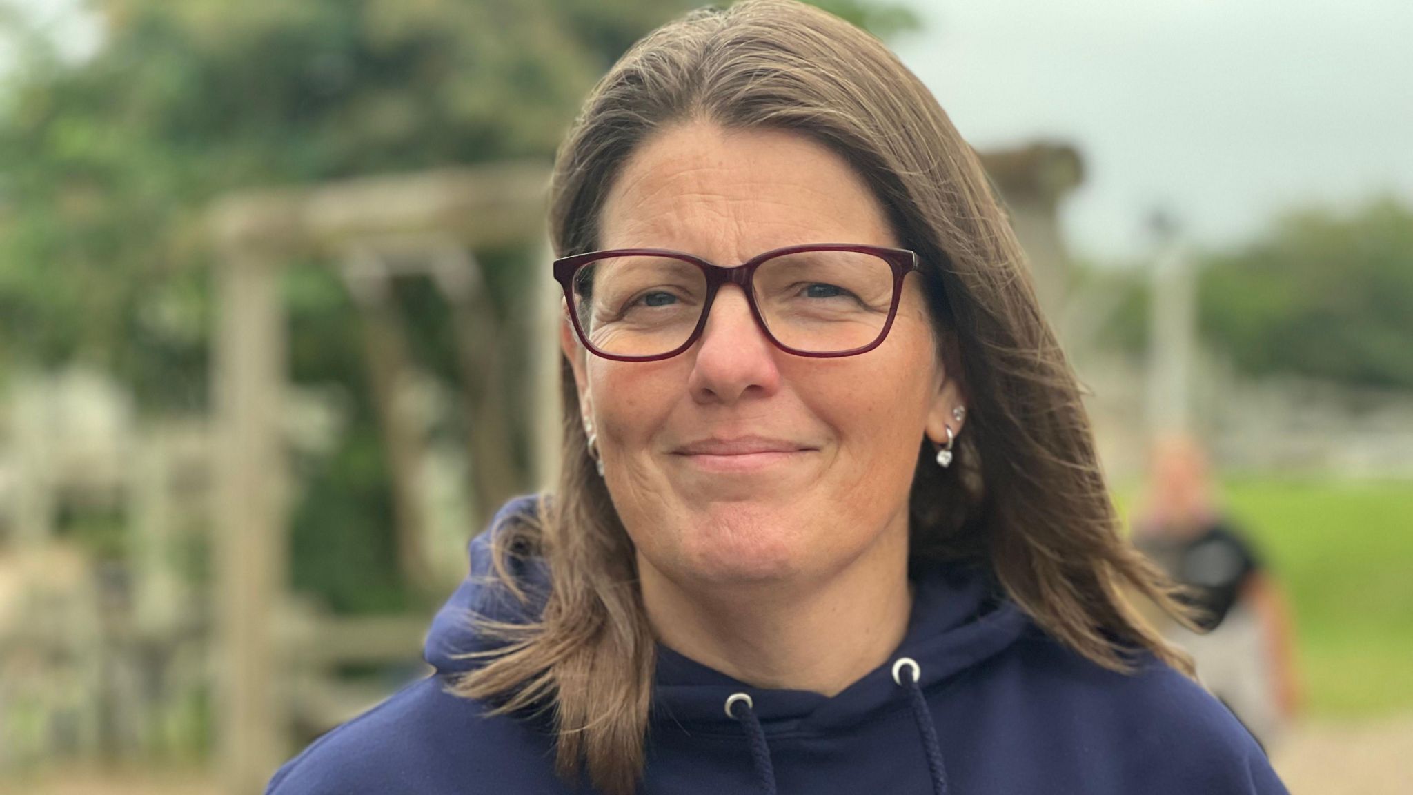 Dawn looking into the camera smiling wearing glasses with mid-length dark brown hair and a navy blue hoodie