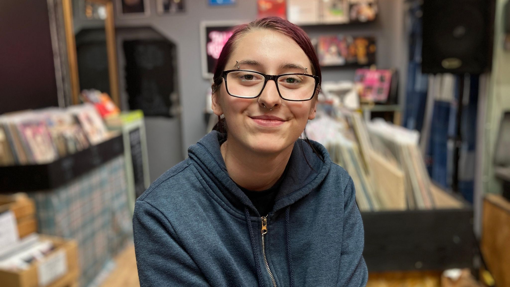 Paris Mehmet is looking straight towards the camera. There are records for sale behind her. She is wearing glasses and smiling