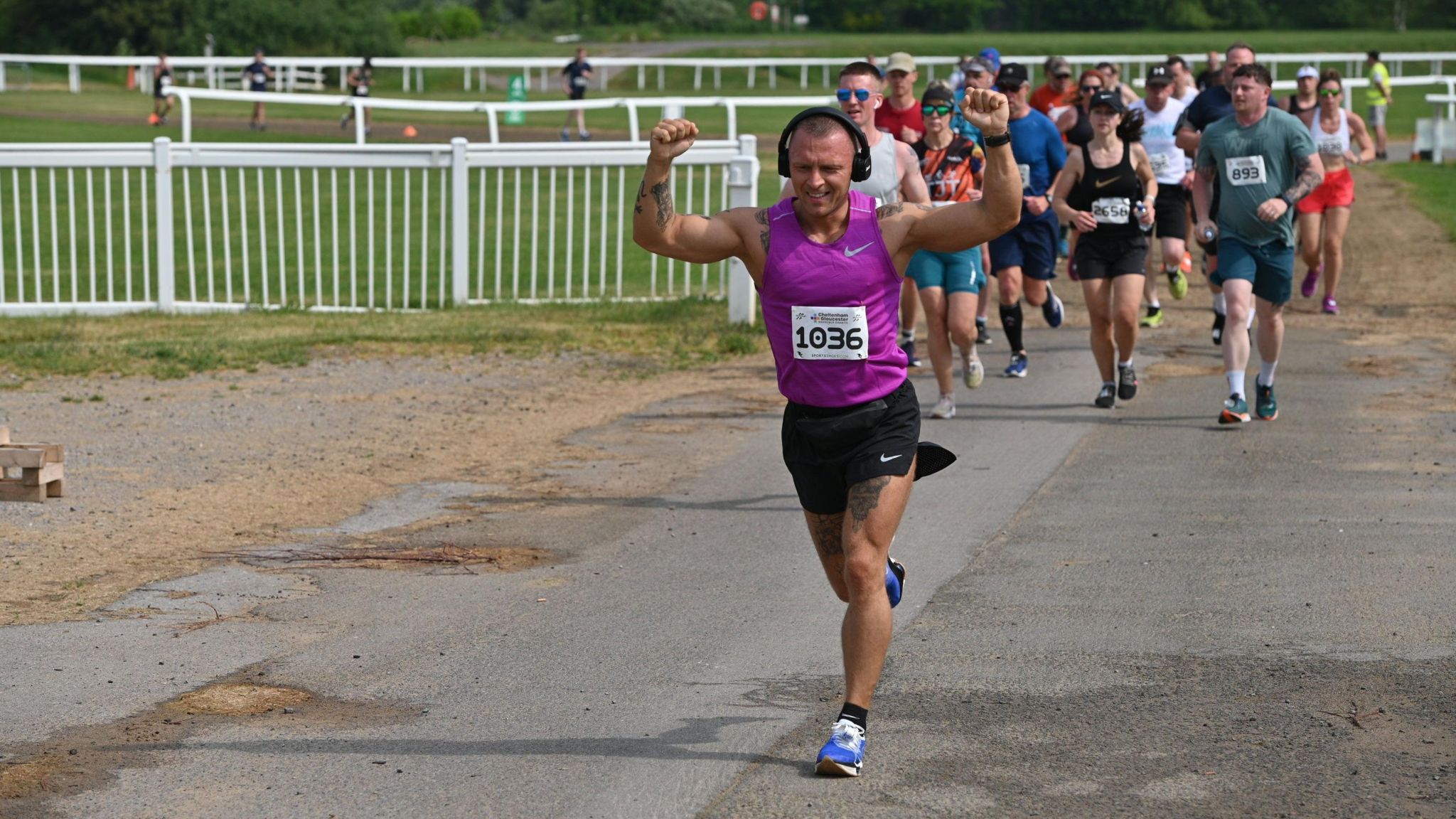 A man on Cheltenham racecourse in running gear holding both arms up while others run behind him