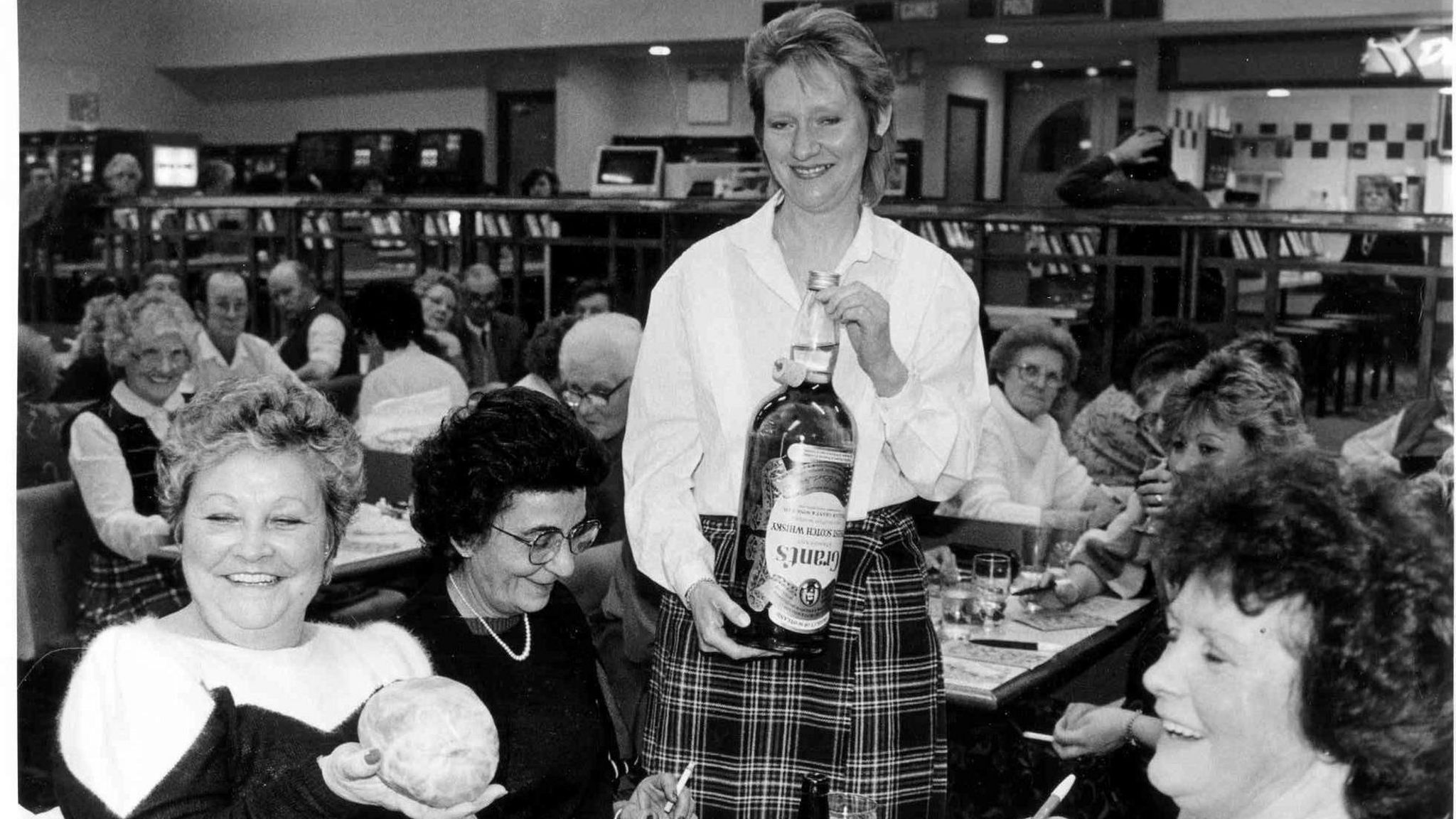 A lady holding a large bottle of whisky presenting it to two seated ladies in the bingo hall, everybody is smiling and laughing