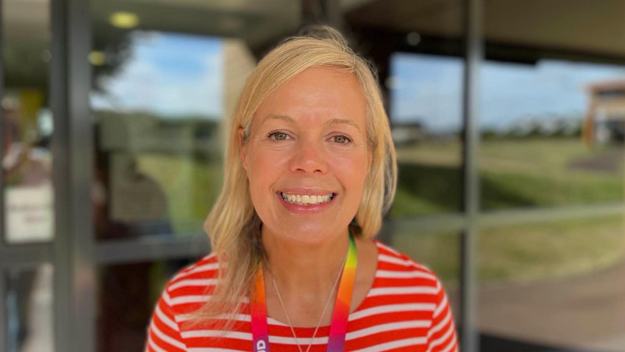 A white woman with blonde hair smiling. She is wearing a stripy red and white shirt with a rainbow lanyard. She is facing the camera and stood in front of windows