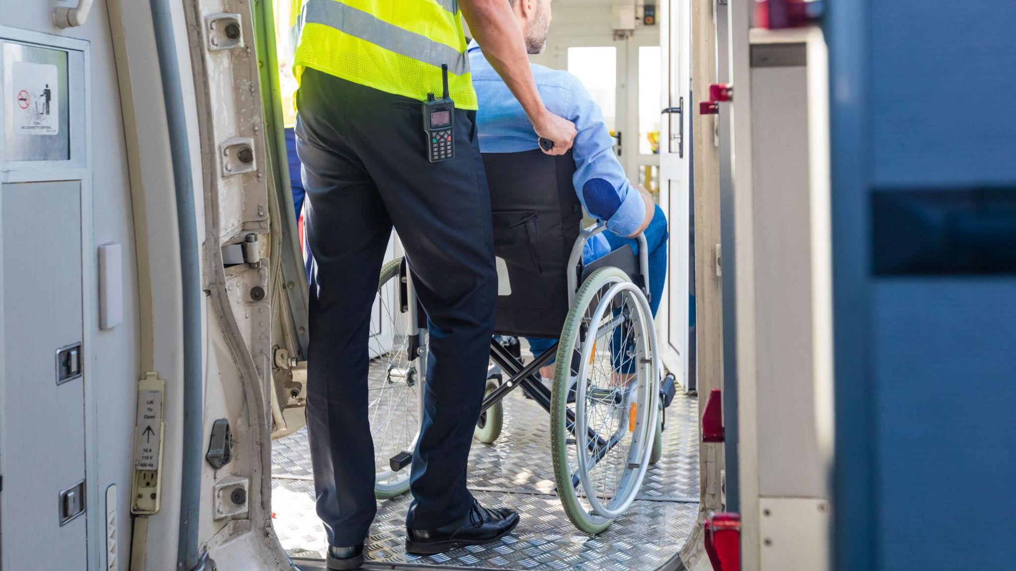 An airline assistance worker wearing a yellow hi-vis vest and a walkie talkie clipped to his waistband. He is pushing a man in a wheelchair through the entrance of a plane. 