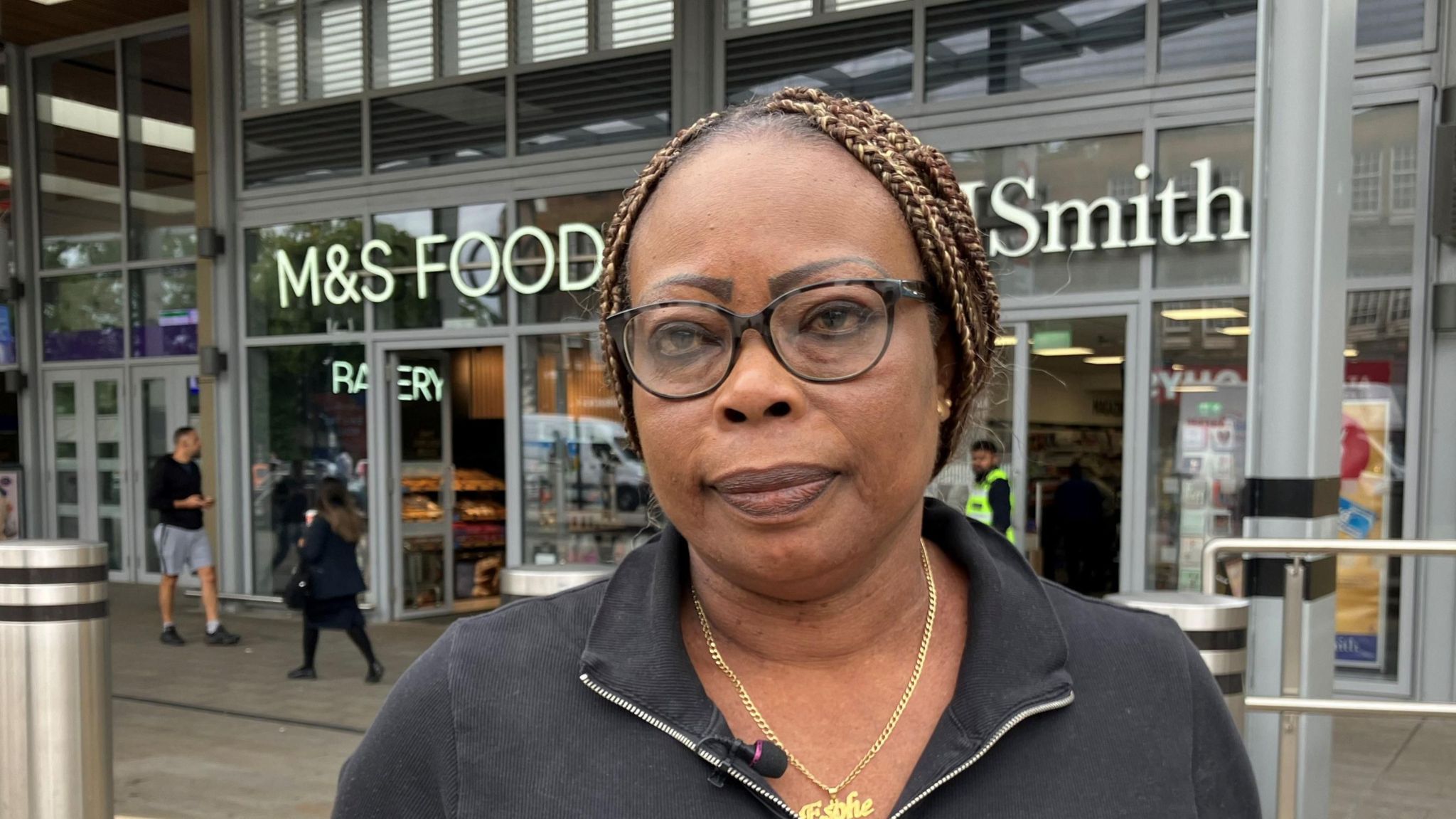 Lucy Daramola standing outside Ealing Broadway station with a Marks and Spencer Food shop and WH Smith in the background. She has light brown braided hair, black rimmed glasses, gold chain necklace and a black zipped top.