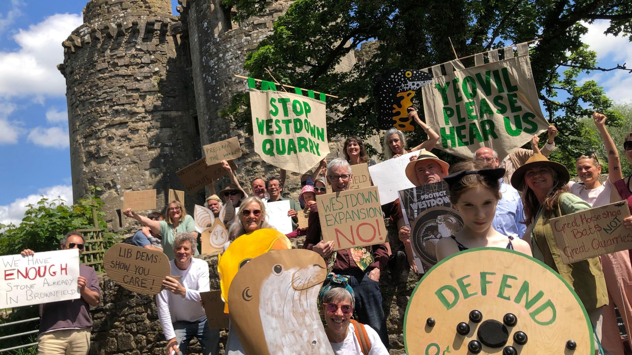 A large group of protestors standing in front of an old castle building. They are all holding cardboard signs or banners that read 'stop Westdown Quarry' and 'Westdown expansion NO!"