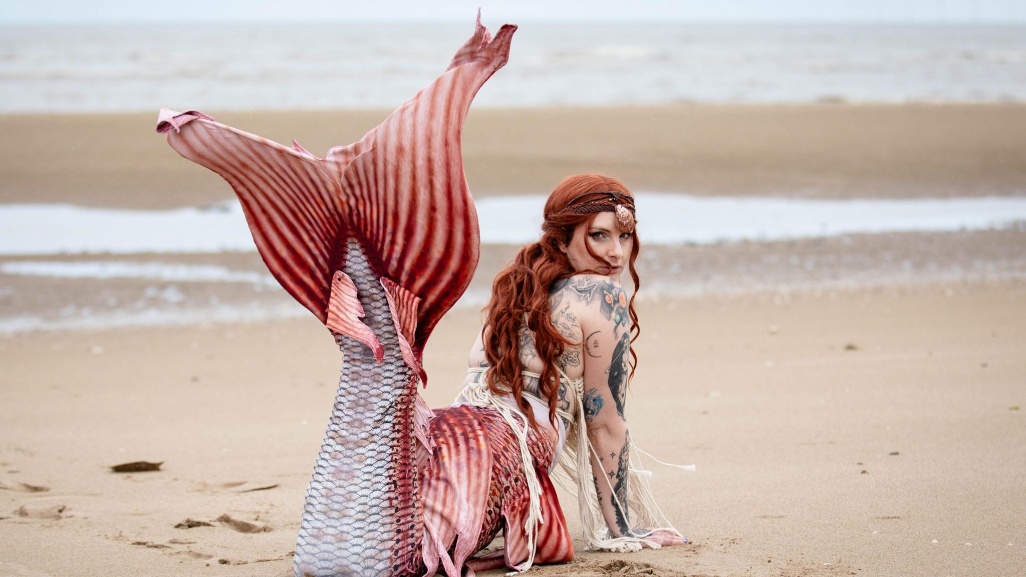 Tyler Turner lying on her front on a sandy beach with a salmon pink, red and white scaled tale on her legs with a matching red wig on her head. The tail is lifted up to the sky and Tyler is looking at the camera. 