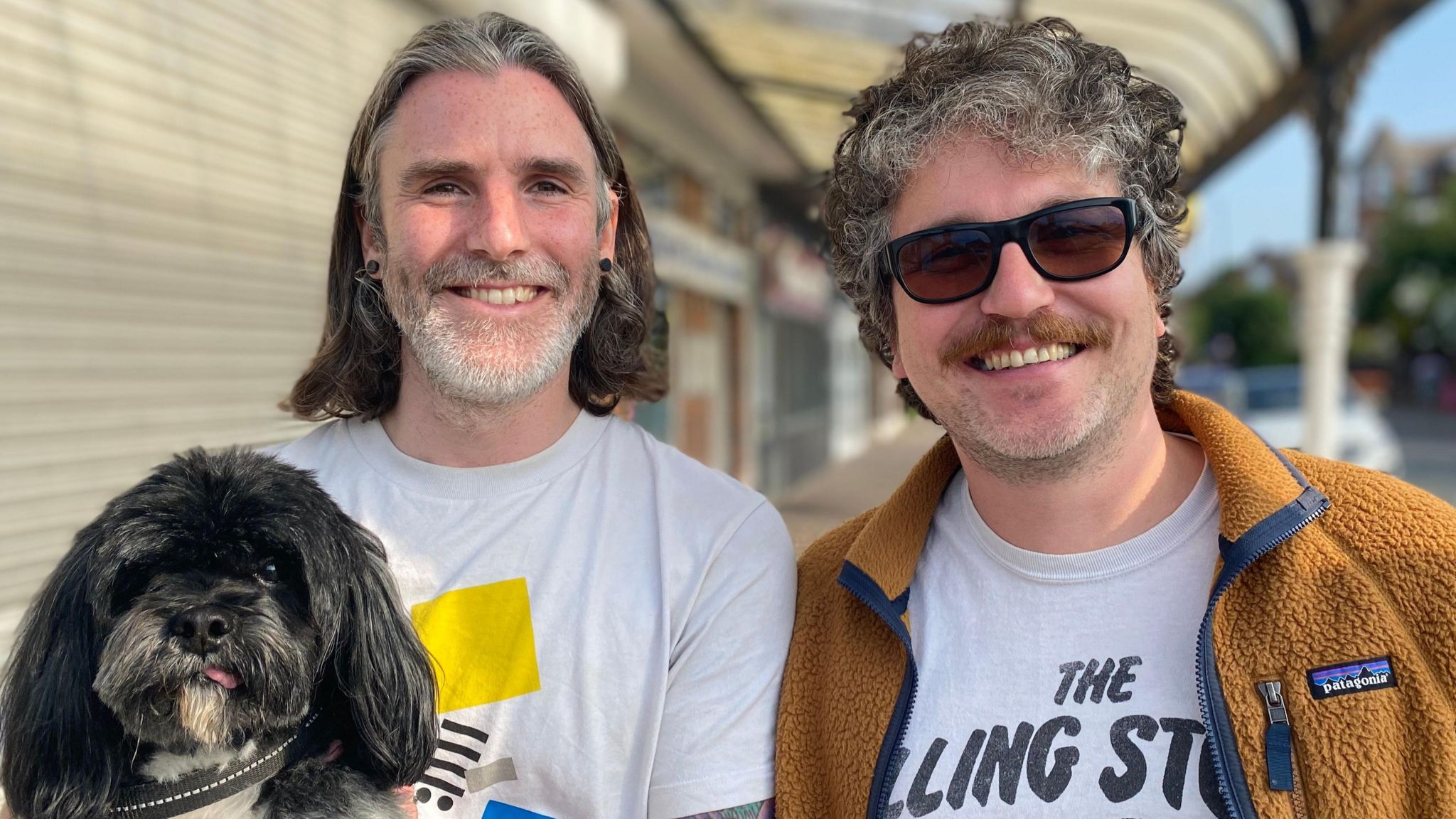 A smiling Steve Smith, with shoulder-length greying hair and a close-cut beard, wearing a white t-shirt with a yellow block design and carrying a small black and grey-haired dog, and a smiling Paul Smith, with curly greying hair, a brown moustache, stubble and sunglasses, wearing a white T-shirt bearing the words 'The Rolling Stones' under a brown fleece jacket, stand in front of a yellowing covered walkway alongside shops, some of which have shutters down