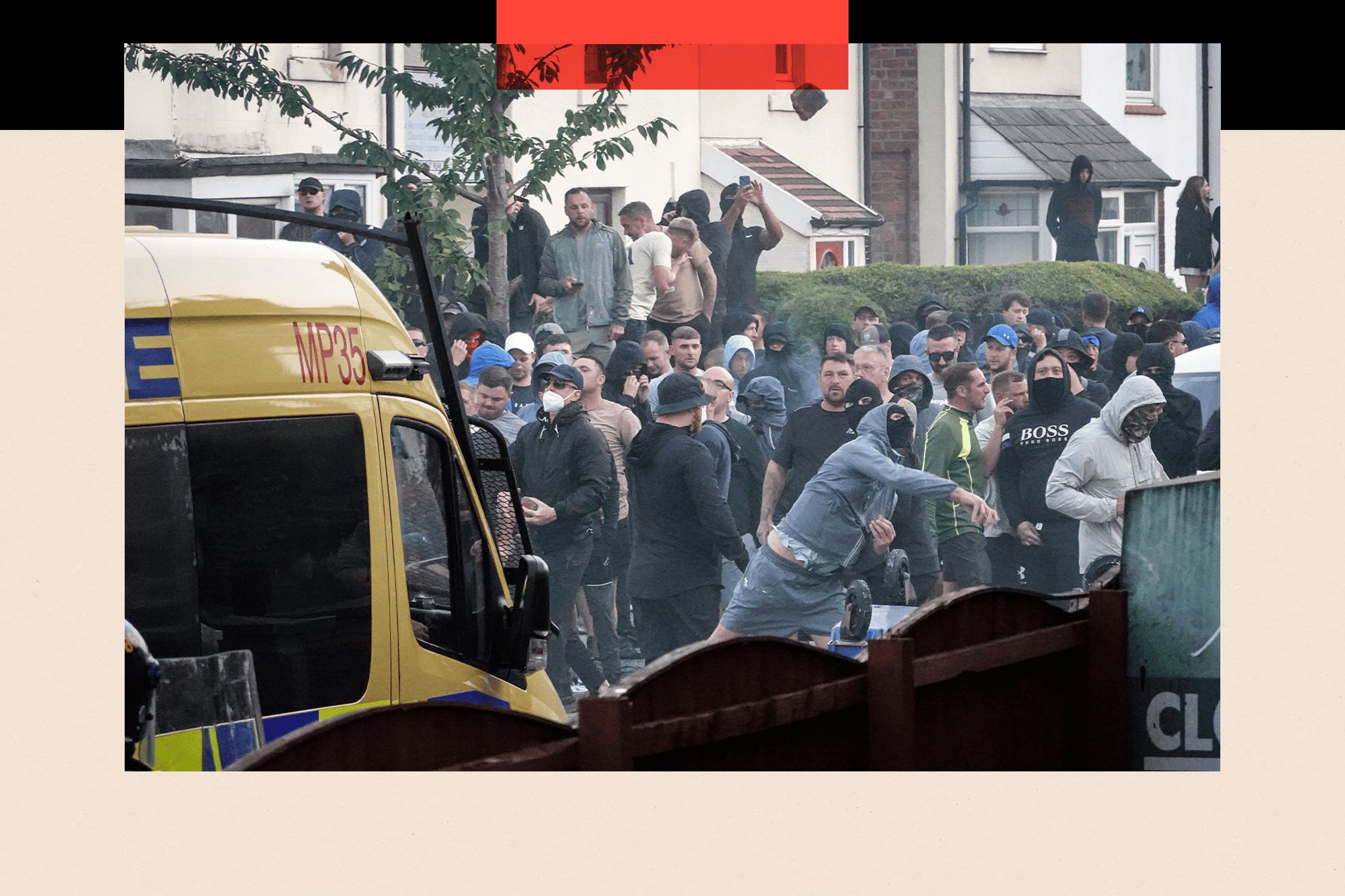 A crowd of male protesters behind a police van. One man in the foreground is wearing a black face covering and blue tracksuit top and shorts. He appears to be hurling something.