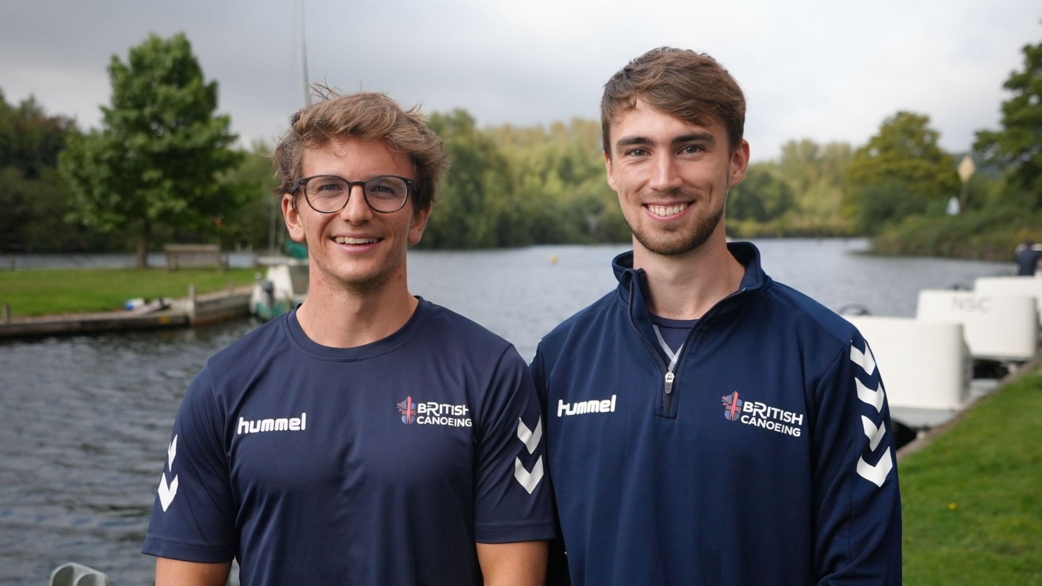 James How and Tim Dowden smile at the camera while standing side by side. How is wearing glasses and a blue T-shirt branded with "British Canoeing" and Dowden is wearing a blue sweatshirt with the same logo. They are standing on a riverbank with the water behind them. 