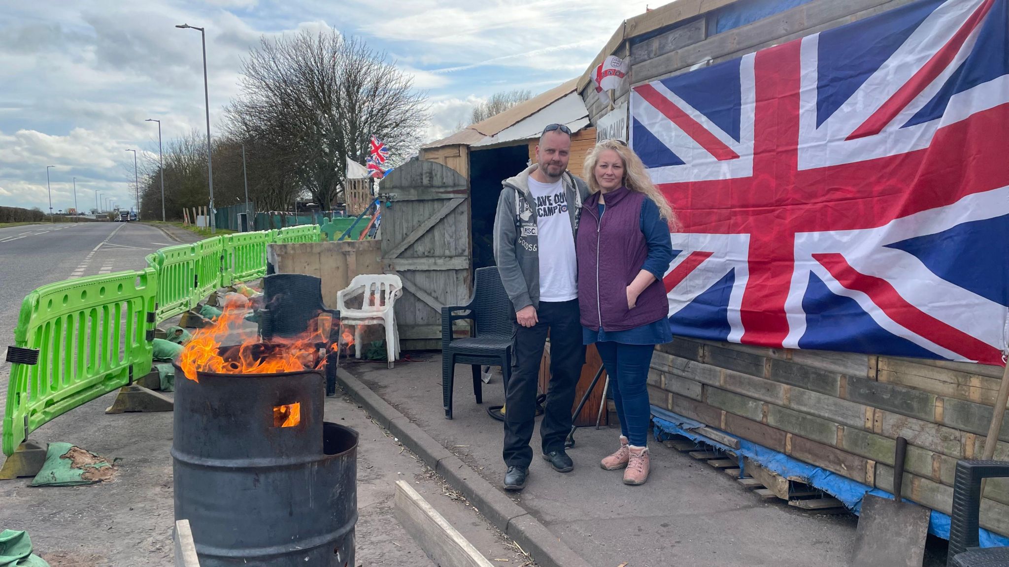 A man and woman stand outside RAF Scampton. A Union Flag is hanging on a wall behind them, a bin has flames coming out of it and there are green barriers on the road.