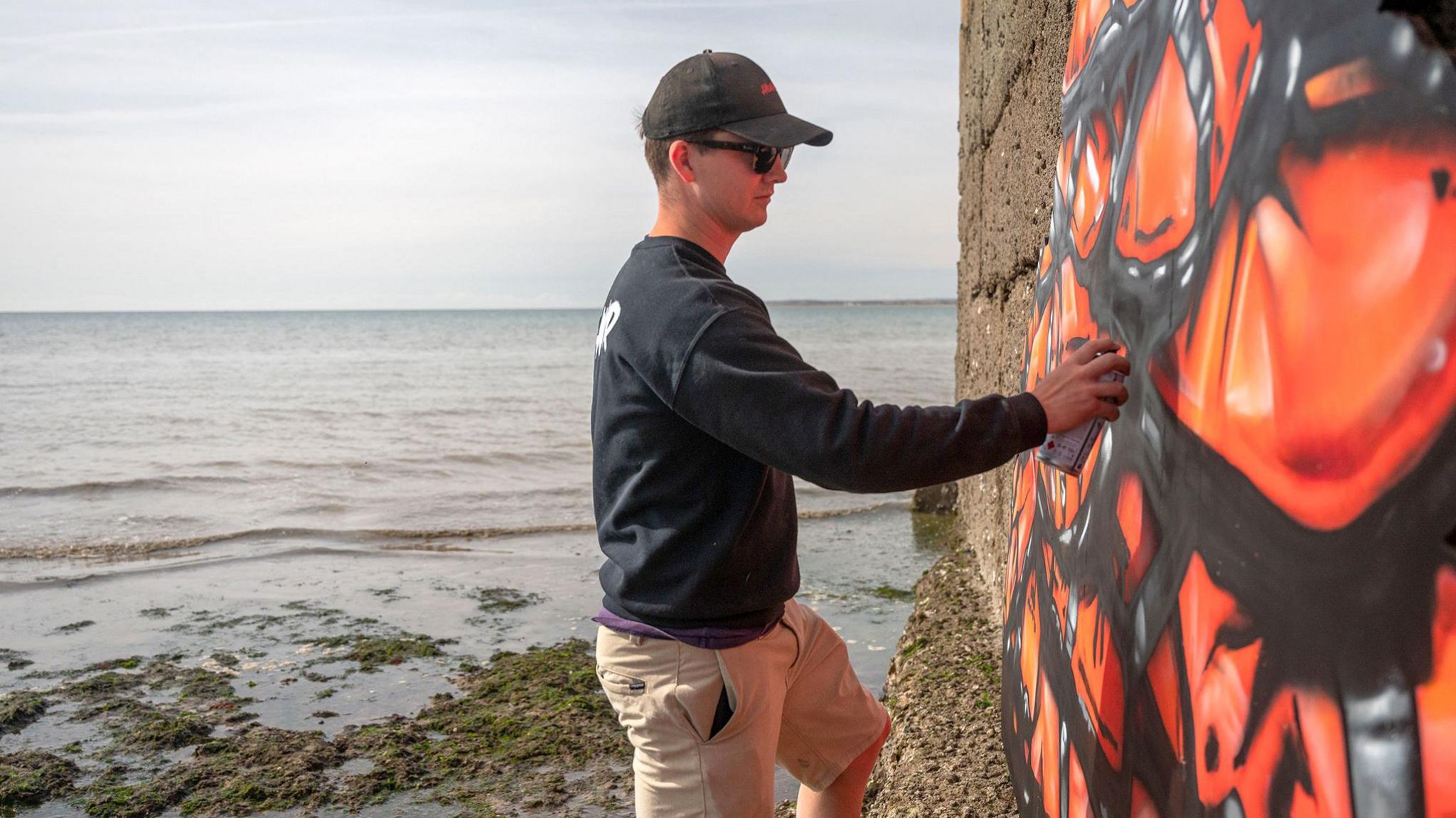 Kent-based artist Hugh Whitaker holds a can of spray paint to the mural in black and orange, with the sea behind him