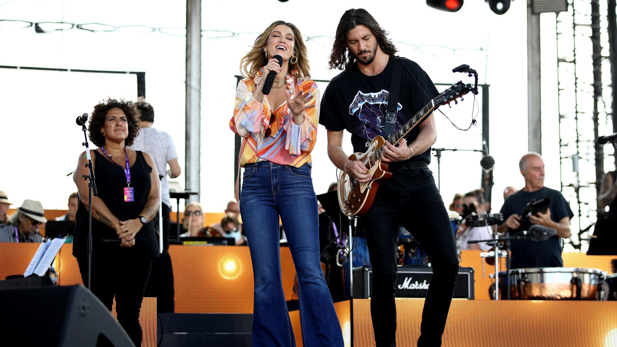 Delta is performing with Matthew Copley. She is wearing a multicolour top and jeans. He is standing next to her in a black t-shirt and black jeans and is playing the guitar. 