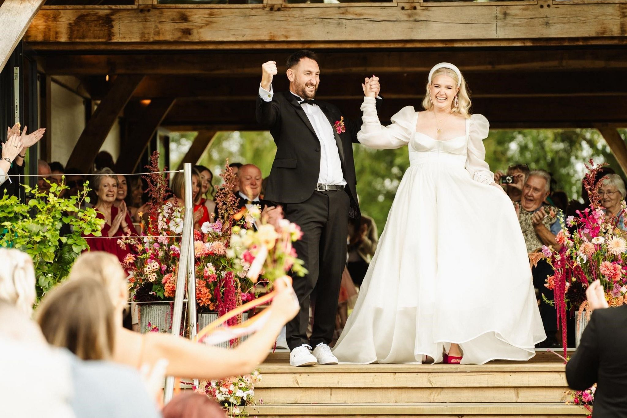 Brooke Jones and her husband Alex are holding hands and celebrating. There are wedding guests seated around them. She is wearing a white wedding dress and he is wearing a black suit with a white shirt