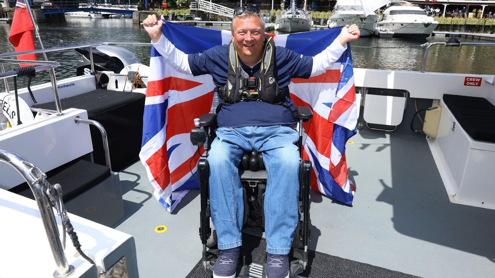 A man in a wheelchair on the back of a boat wearing a life jacket smiling and holding up a union jack