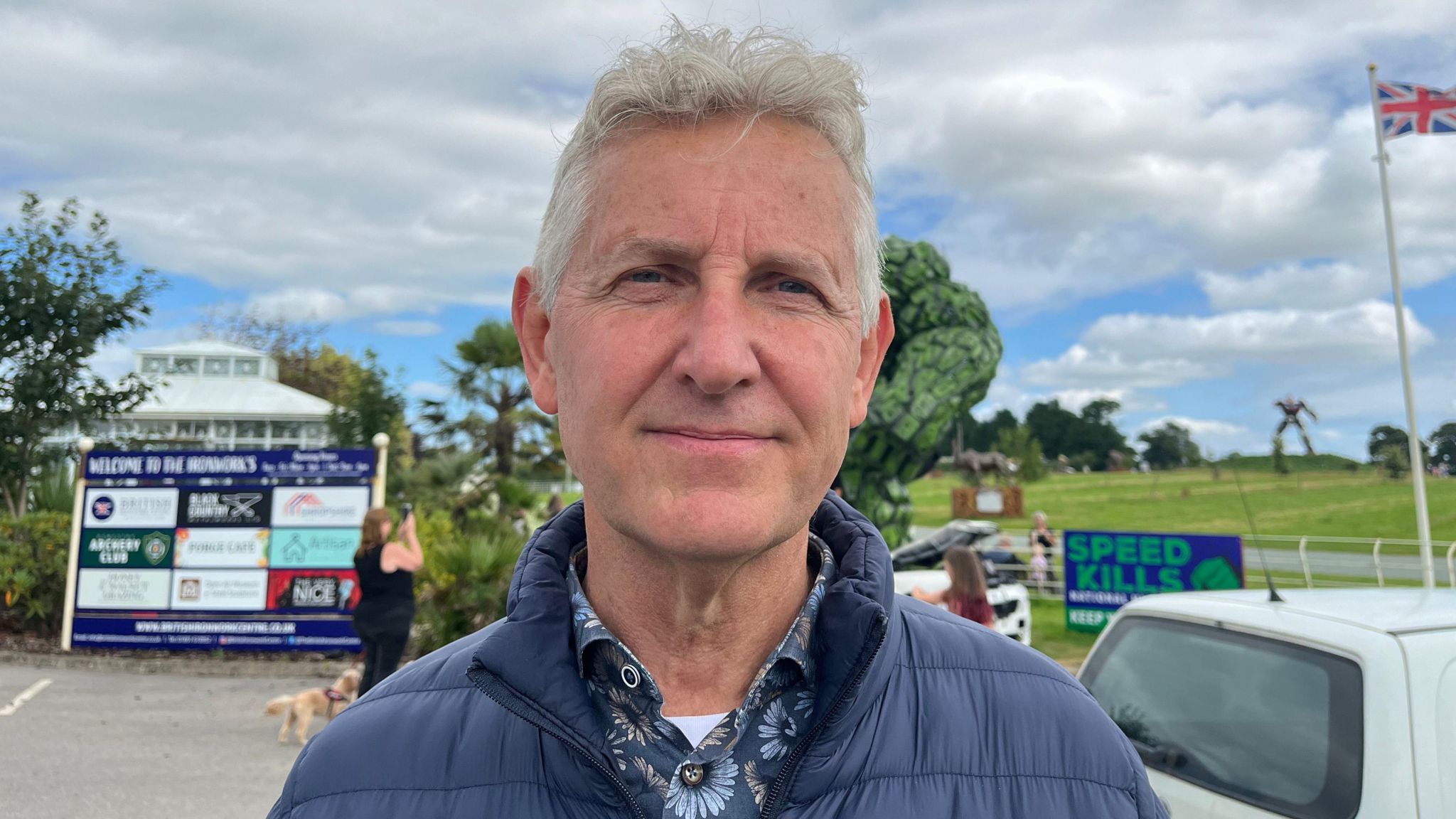 Clive Knowles is smiling into the camera. He is wearing a dark blue shirt with blue flowers on it, and over the top is wearing a navy jacket. He has grey hair. In the background is a cloudy blue sky and green grass. There is a white flagpole with the union jack flying at the top.