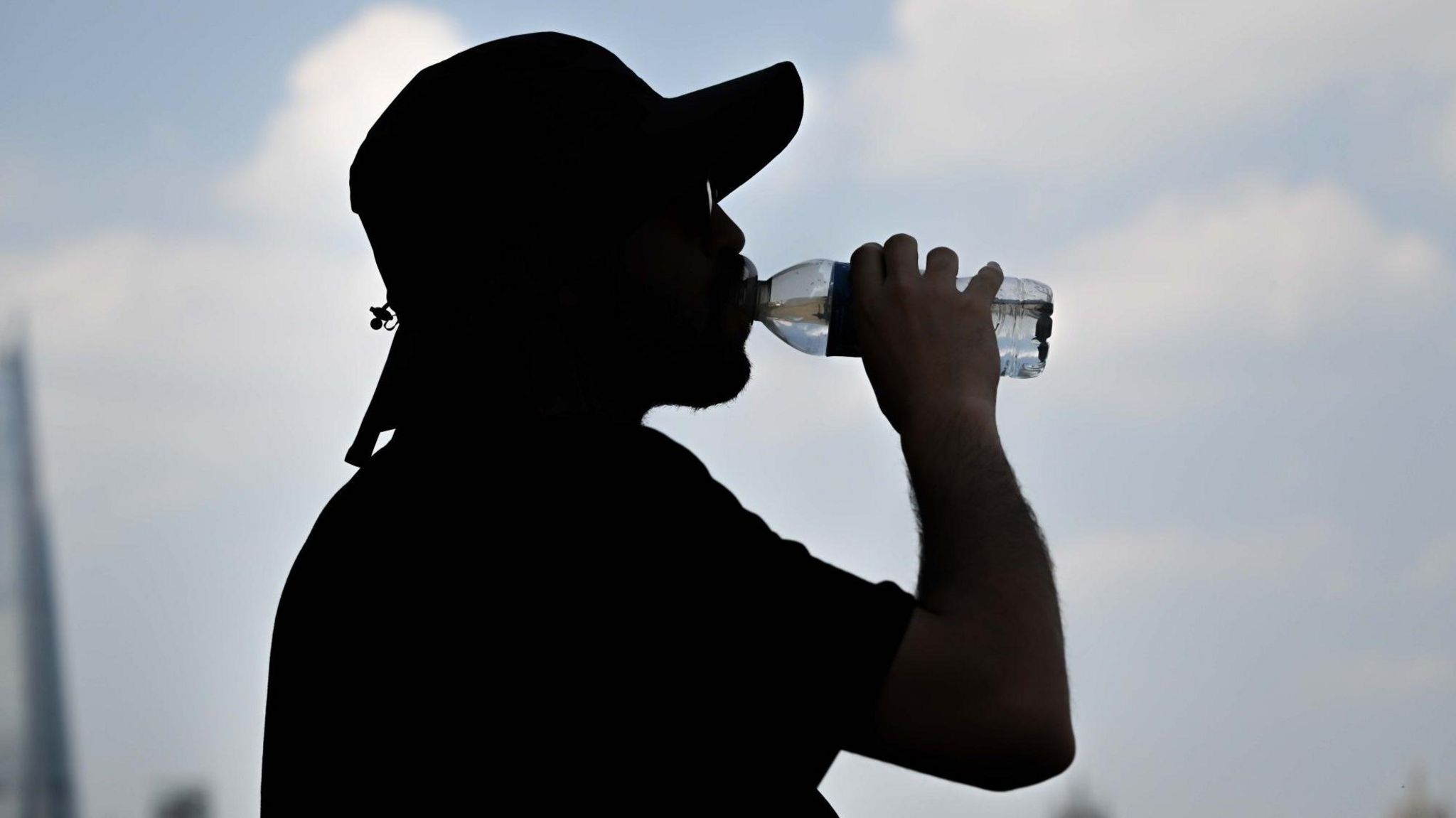 The head and shoulders of a man in silhouette drinking from a bottle of water on a hot day. He is wearing a cap and sunglasses against a blue sky.