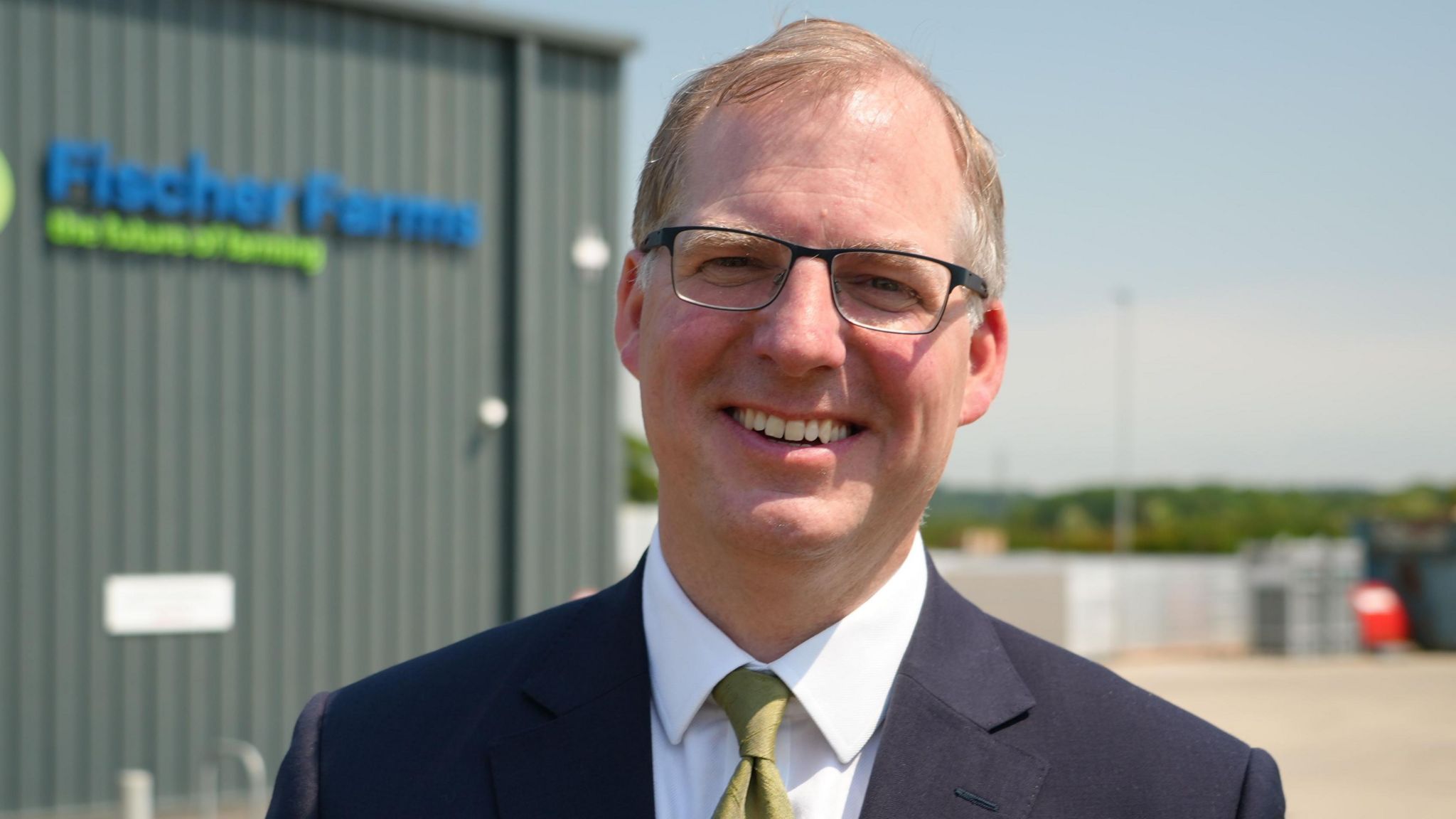 A man in a dark suit with a white shirt and green tie and wearing glasses outside a commercial unit bearing a sign that reads "Fischer Farms"