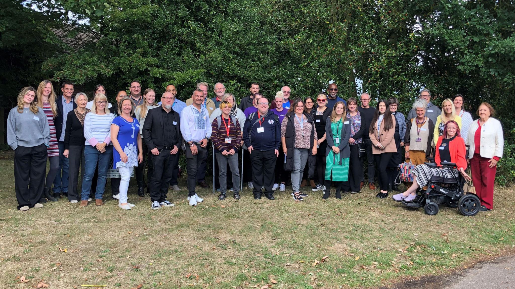 About 40 people stand for a group photo in front of some trees, they are all smiling at the camera. 