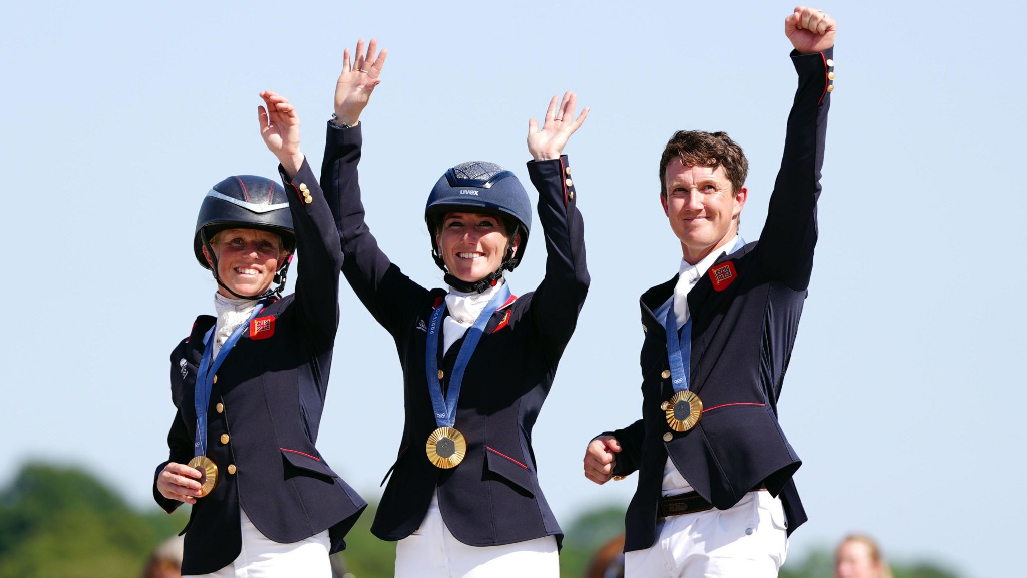 Great Britain's Rosalind Canter, Laura Collett and Tom McEwen with their gold medals. The trio all have their arms up. Tom is holding his fist up to celebrate. They are all wearing equestrian gear and gold medals. 