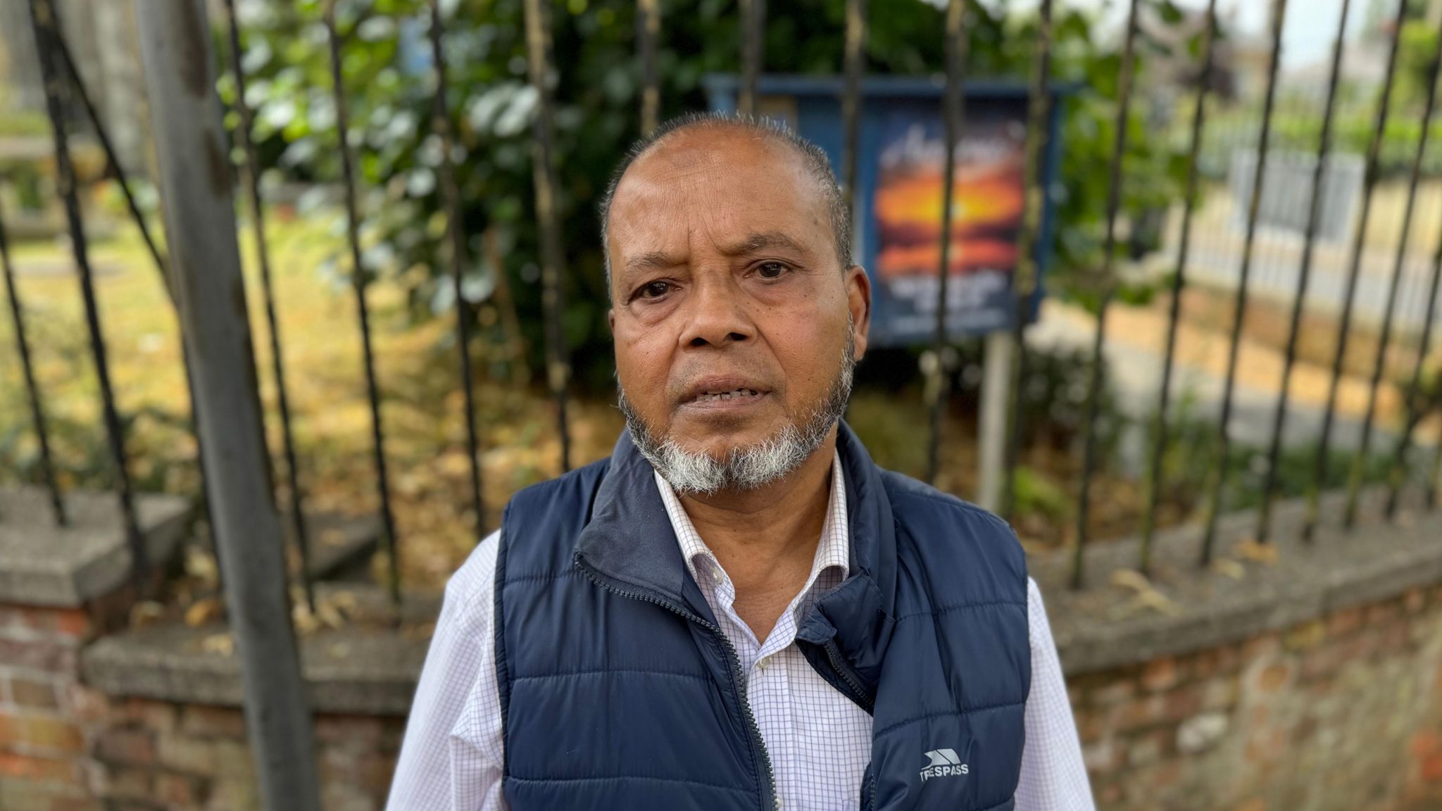 Kamal Uddin looking at the camera wearing a shirt and gilet, standing in front of a stone wall and railings