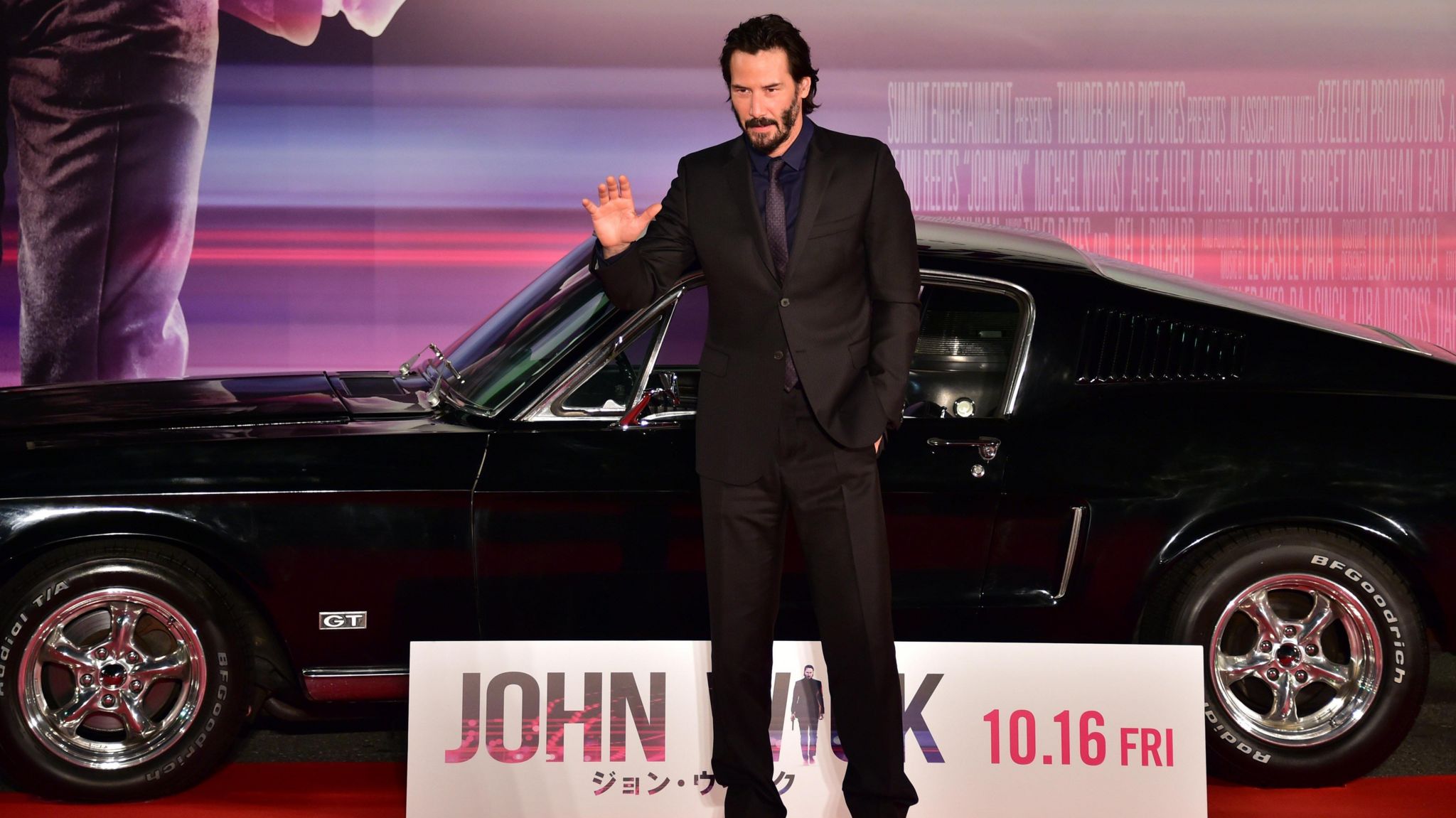 Keanu Reeves, wearing a black suit, at a John Wick premiere in front of a black sports car