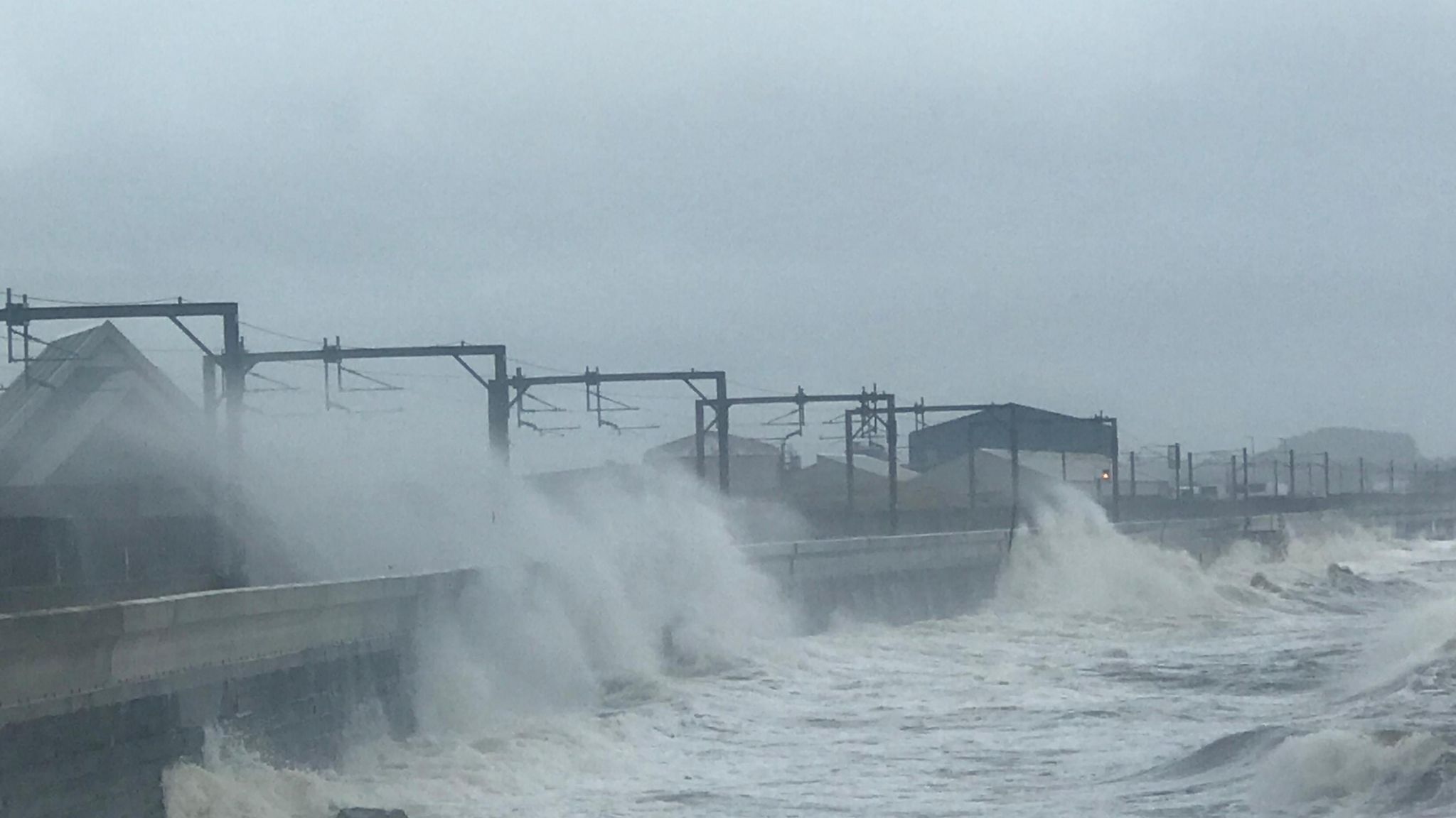 very windy conditions on a coastal stretch of land with waves crashing over a concrete wall onto a railway line.  image is very grey.