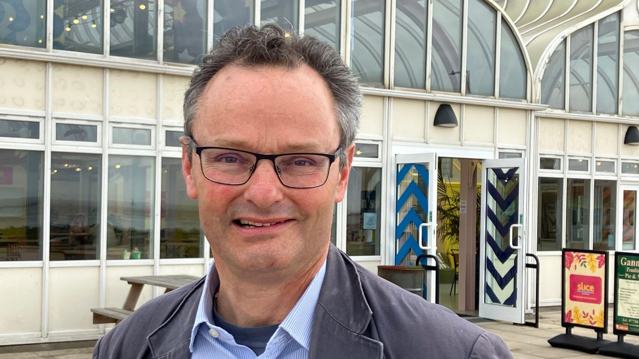 The Conservative candidate wearing glasses and a jacket stands in front East Point Pavilion