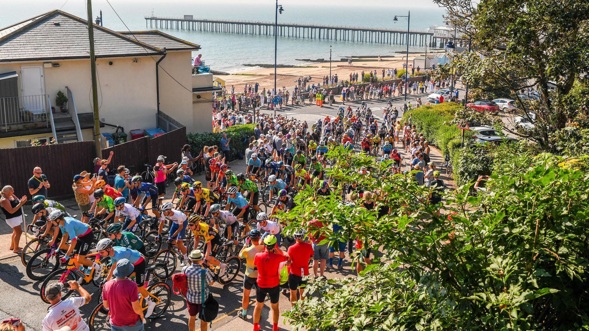 A large group of cyclists in helmets and spandex line up at the start line, with spectators taking photographs and cheering them on from the side.