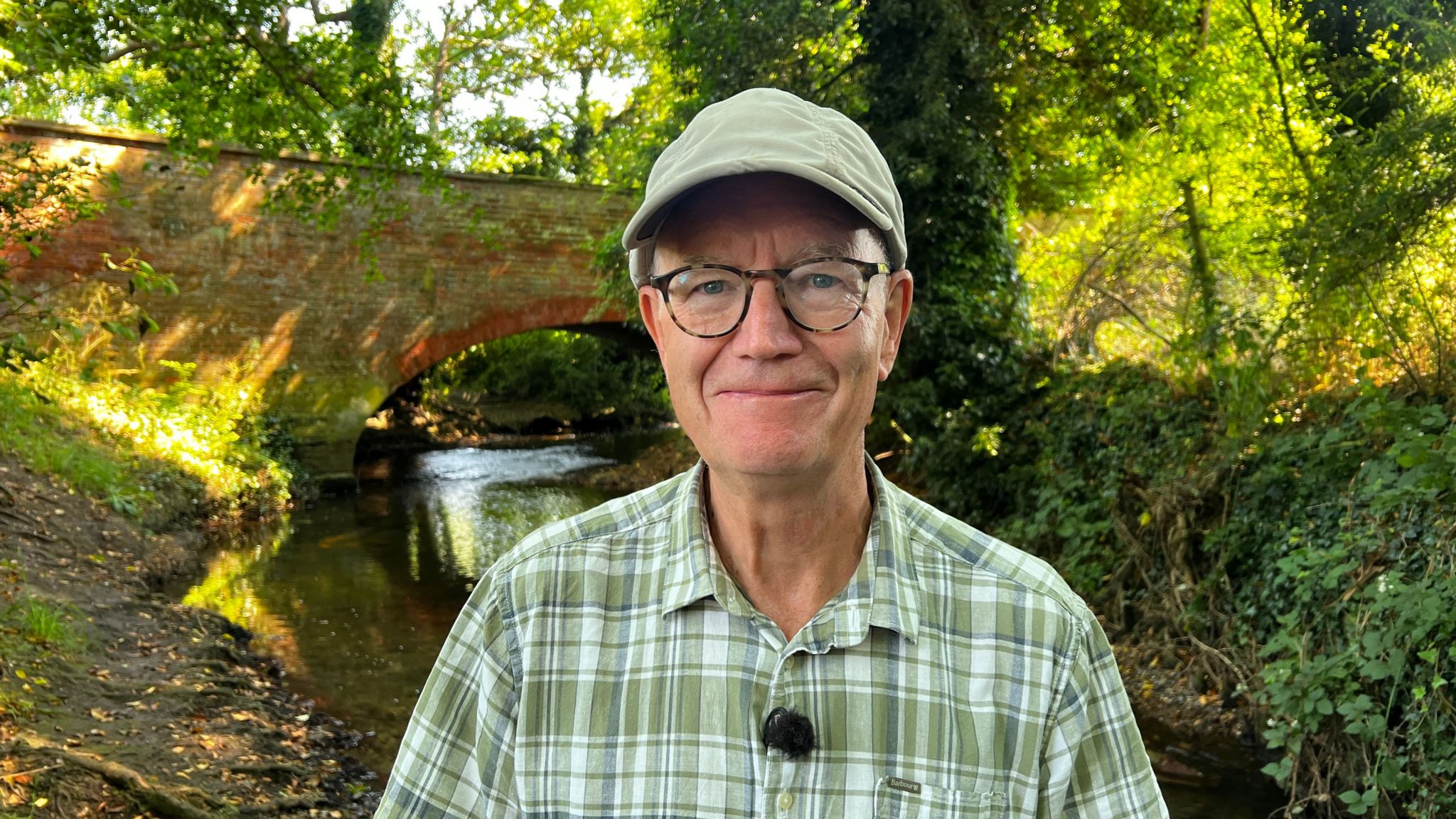 Man wearing a green plaid shirt and standing in front of a sun dappled river surrounded by trees and with a stone bridge spanning it behind him