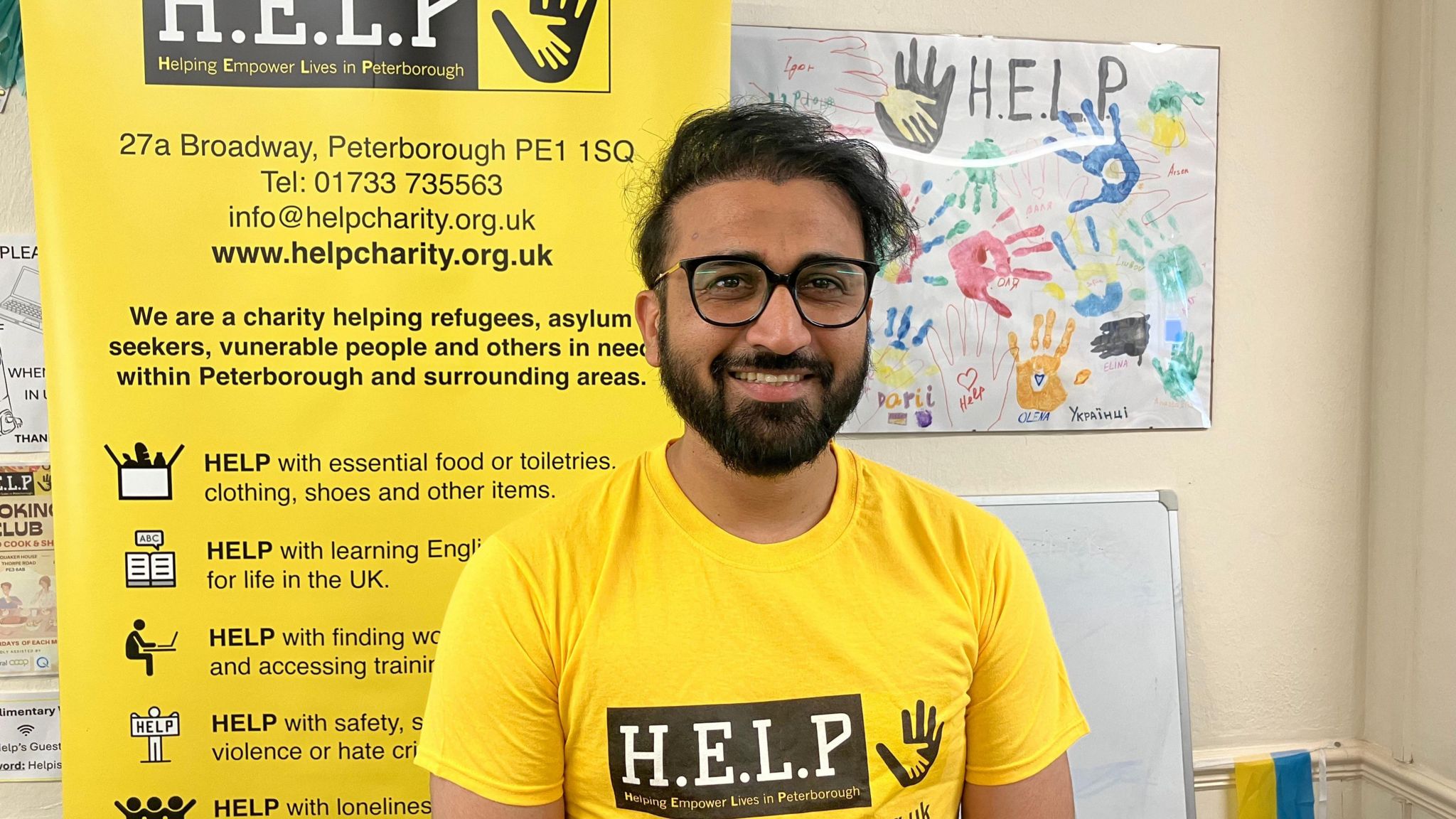 Smiling at the camera is Bilal Aslam, who is wearing a black circle rimmed glasses and a bright yellow t-shirt with the words H.E.L.P. printed on a black text box. He has black hair, a black beard and is posing for the photo 
inside an office. Behind him is an advert for the charity.