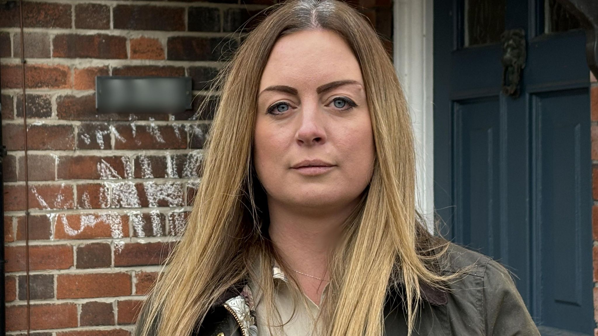 Claire Massey standing outside her home that was damaged by fire in Stoke-on-Trent. She has long light hair past her shoulders and wears a dark jacket over a light blouse. Behind her is a blue front door next to a bare brick wall with graffiti on it