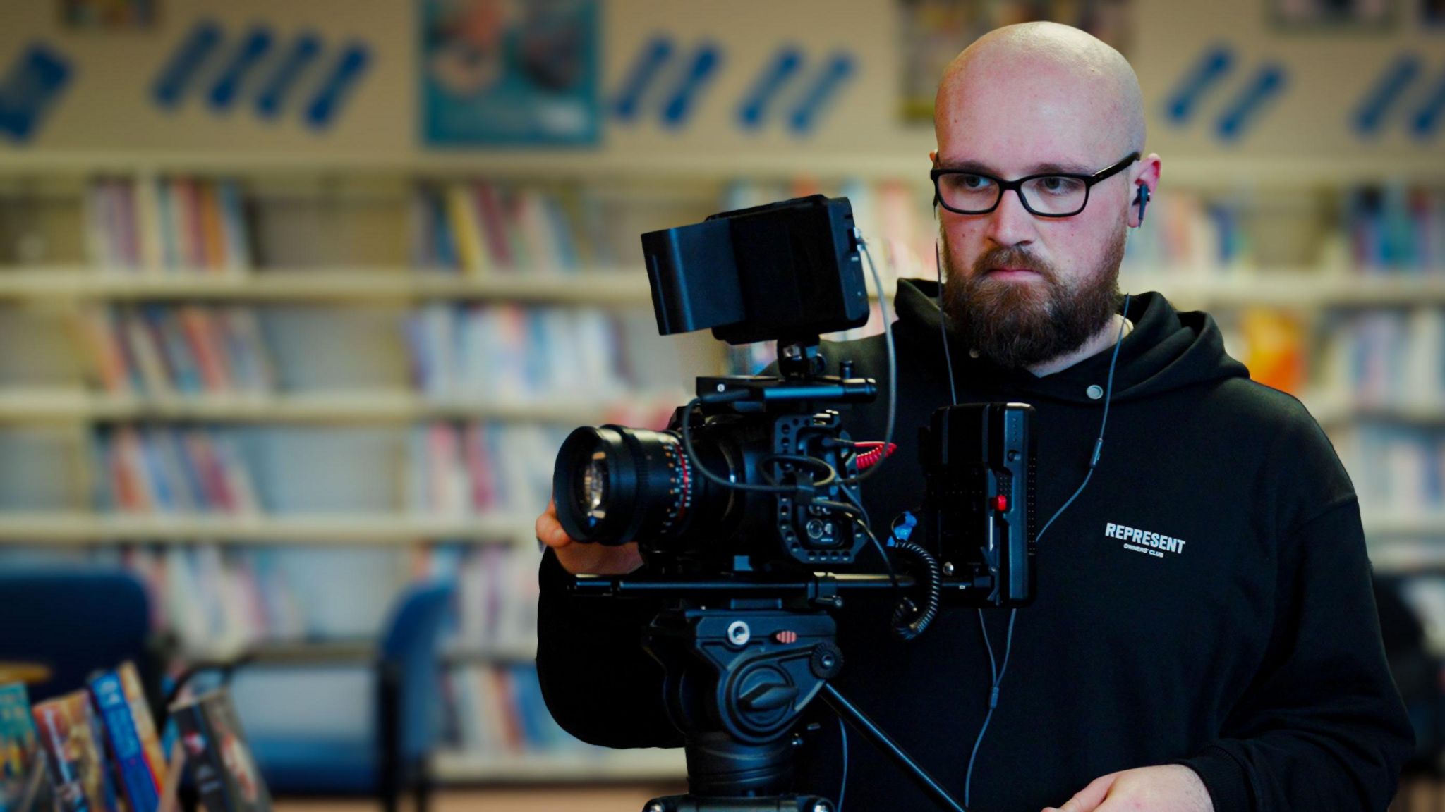 Thomas is adjusting a black professional camera. He's wearing dark rimmed glasses and a black hoody. The blurred background shows library shelves.