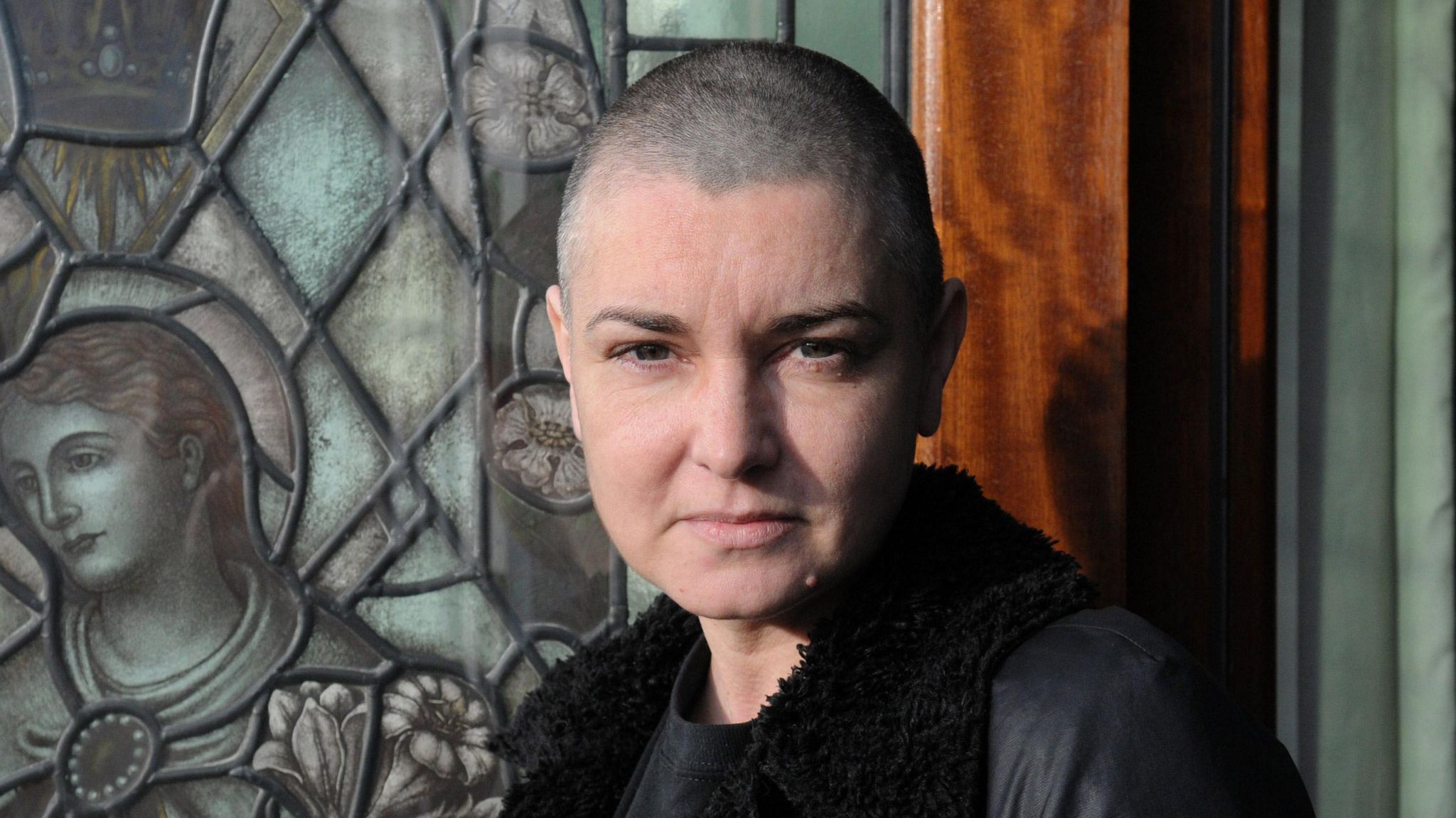 Sinéad O'Connor - A woman with a shaved head looks at the camera with a closed-mouth smile in front of a stained glass window portraying religious imagery