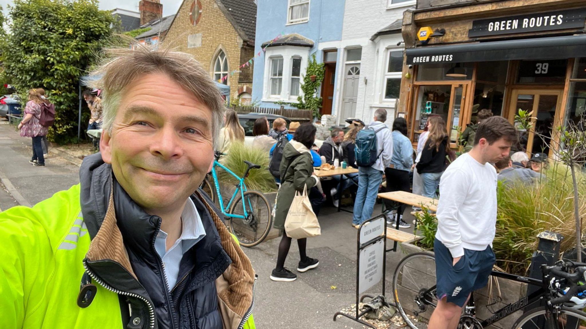 Andrew Gant at the food festival. He is in the foreground wearing a florescent yellow jacket over a black puffer jacket and pale blue shirt. He has short, grey hair. There are several people sitting at a table outside a cafe in the background