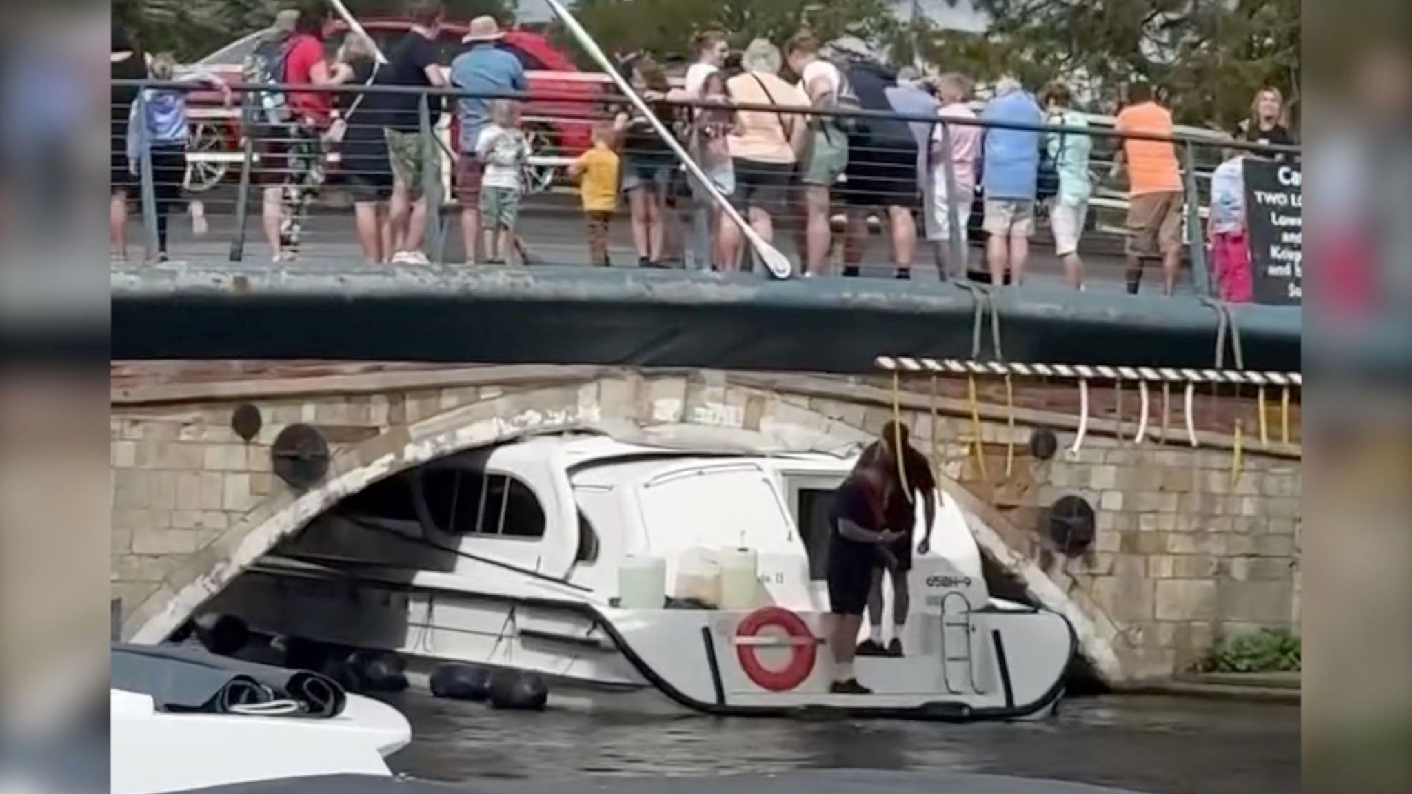 A white boat stuck under a low bridge with a crowd standing on the bridge, some of whom are looking down at the boat. 