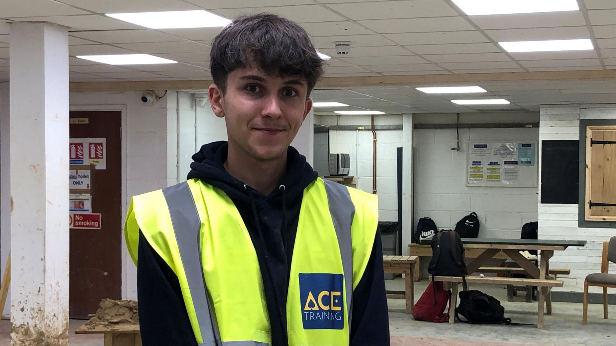 Remus wearing a high-vis jacket and a black hoodie is standing in a white workshop with benches in the background. There is a pillar with chipped paint behind his shoulder and there are six rucksacks on one of the benches. 