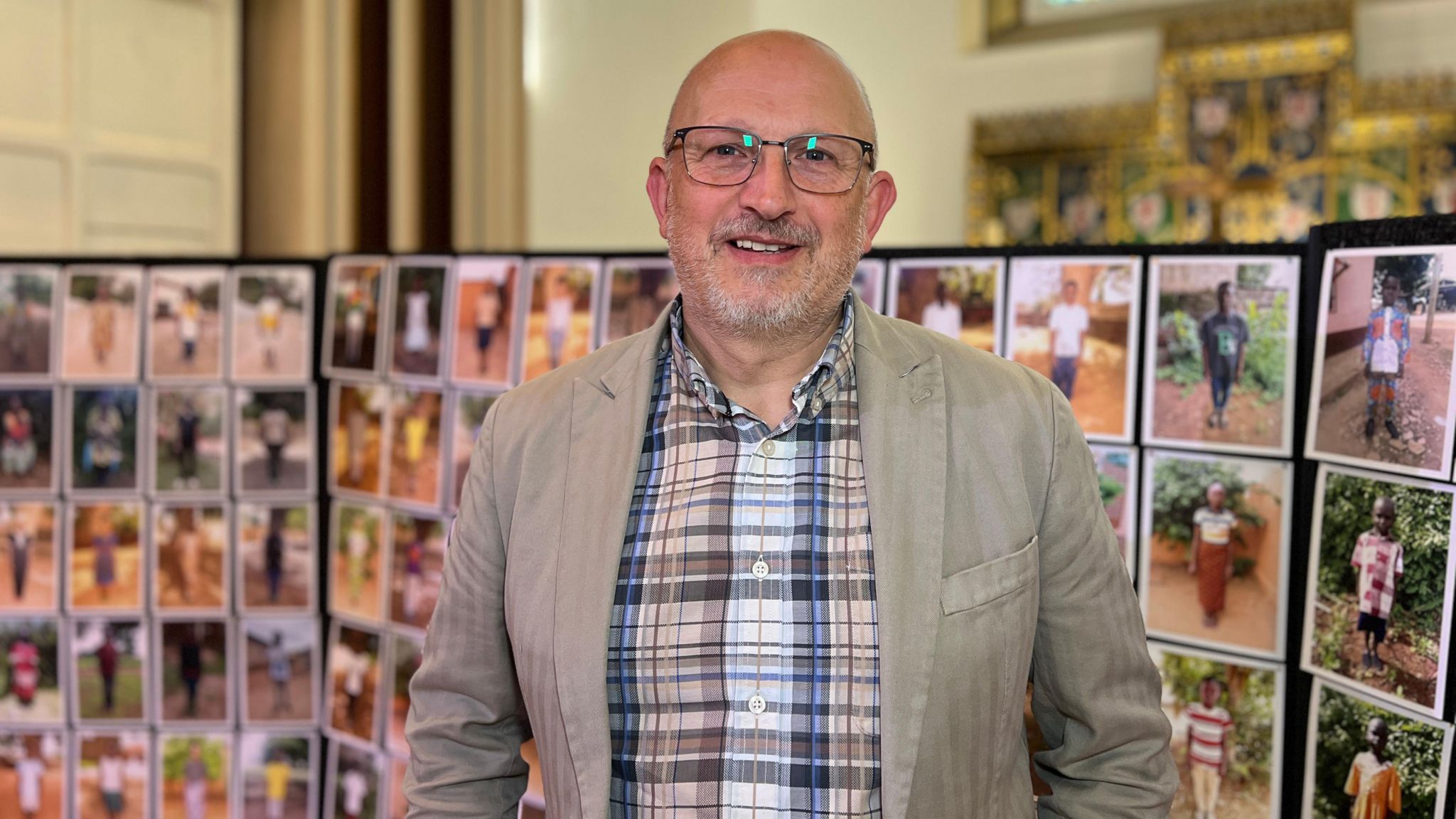 Man smiles at the camera as he stands in front of photos of people supported by Compassion UK