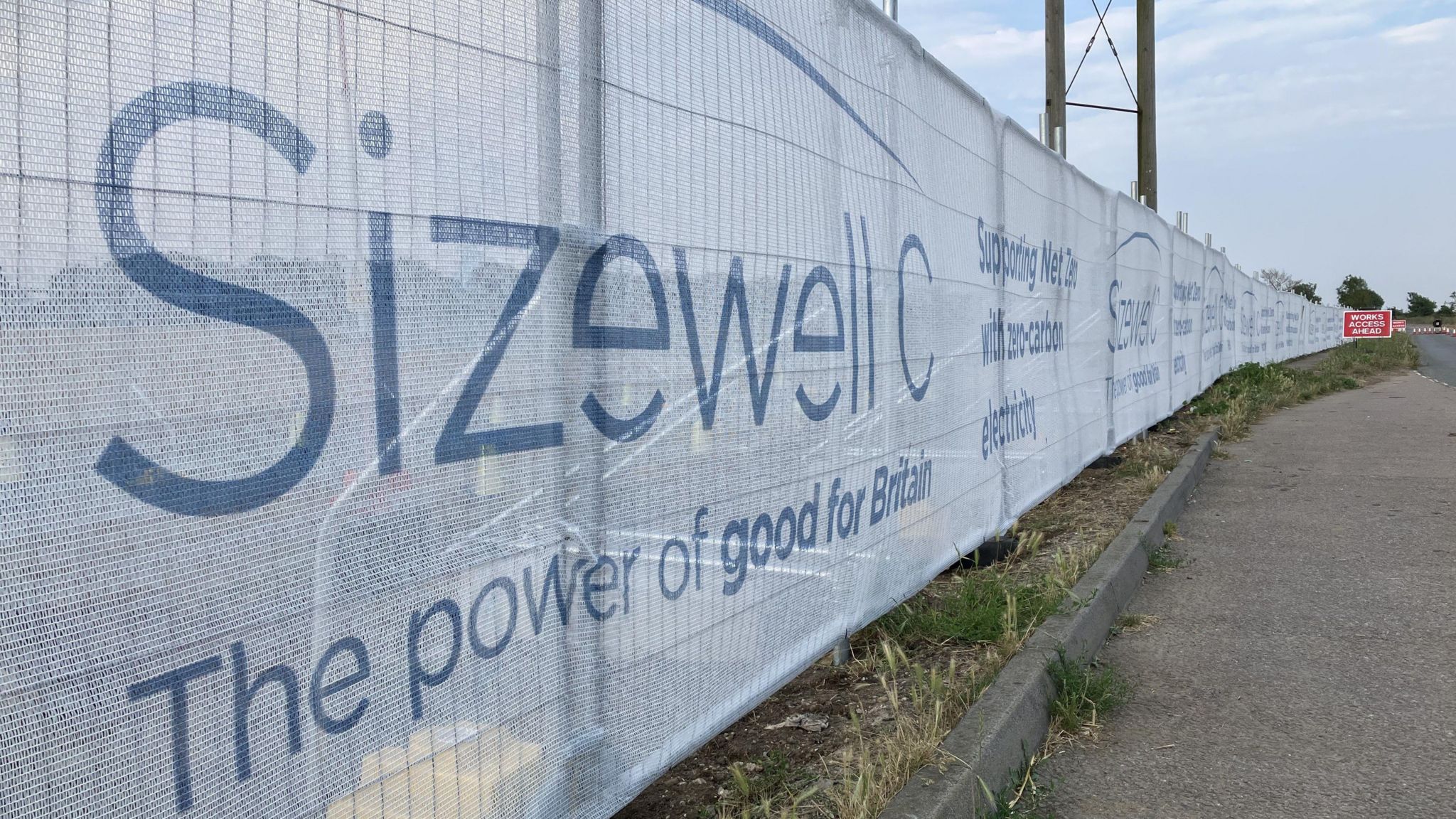 A long metal fence covered in white material with the logo "Sizewell C - The Power of good for Britain'' on it  