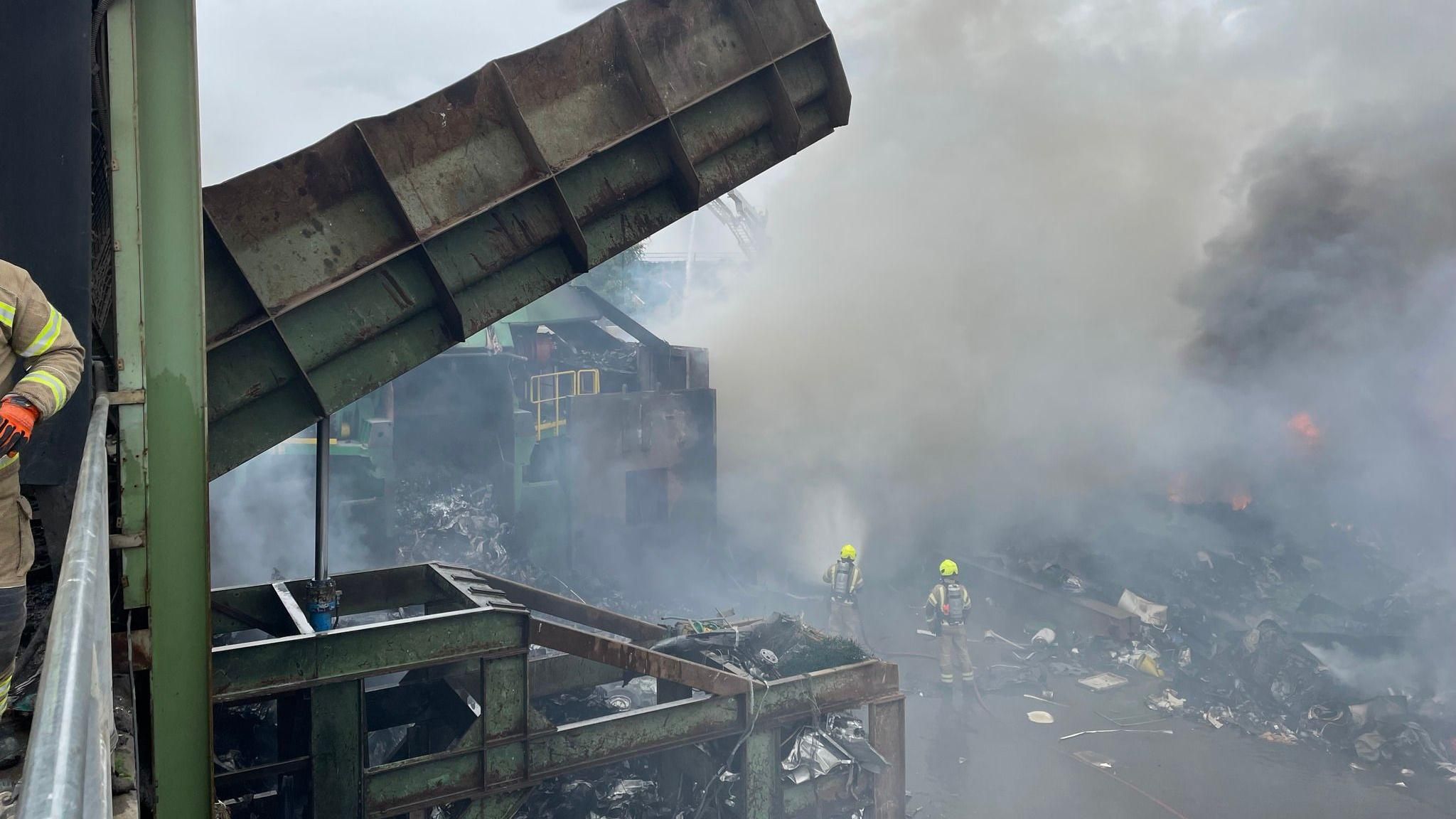 Firefighters at the scene of a recycling centre fire after it has been extinguished. Lots of smoke and firefighters in protective gear damping down. Lots of metal structures in foreground.