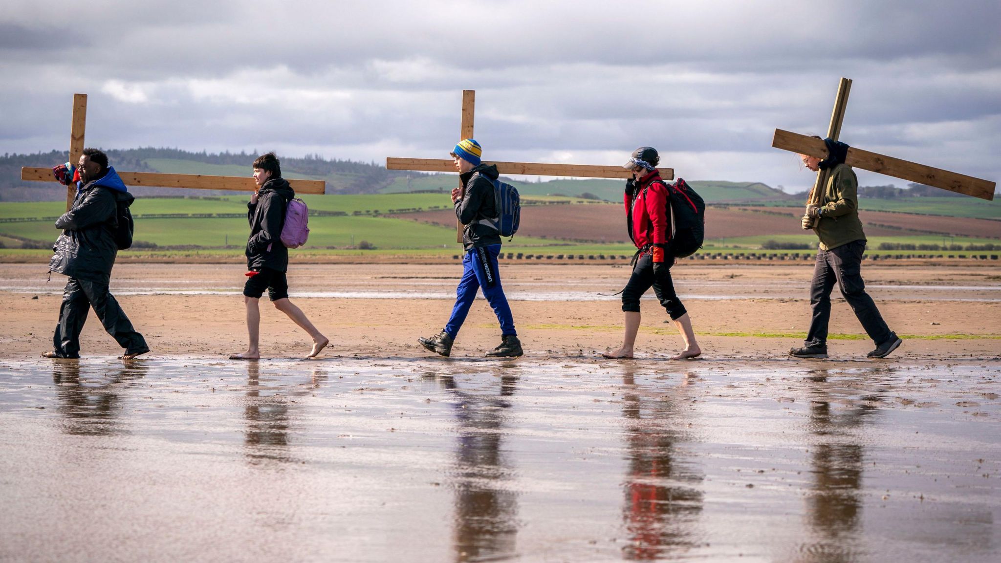A group of five people carrying wooden crosses across the sand