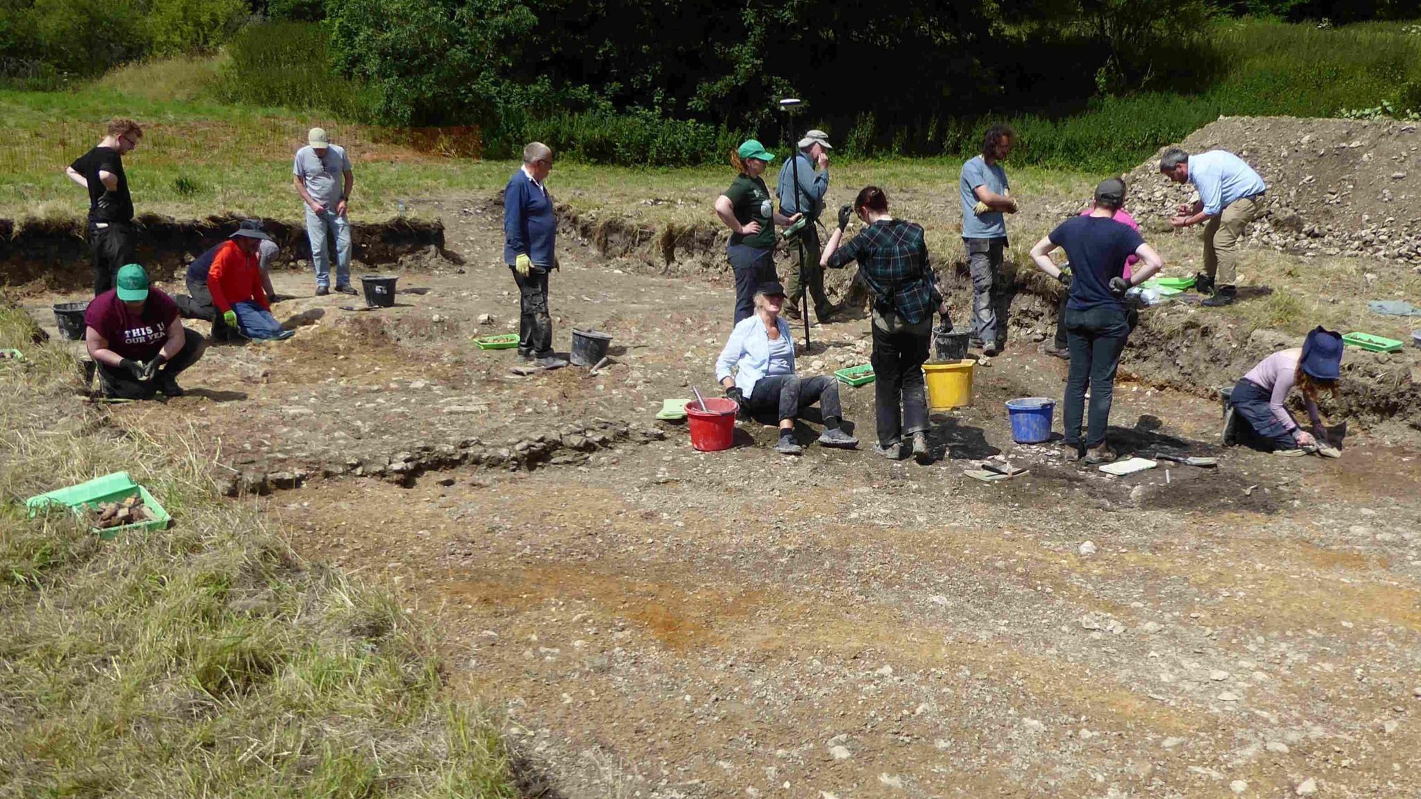 Thirteen people working on an archaeological trench. The remains of a wall can be seen across the centre of the trench