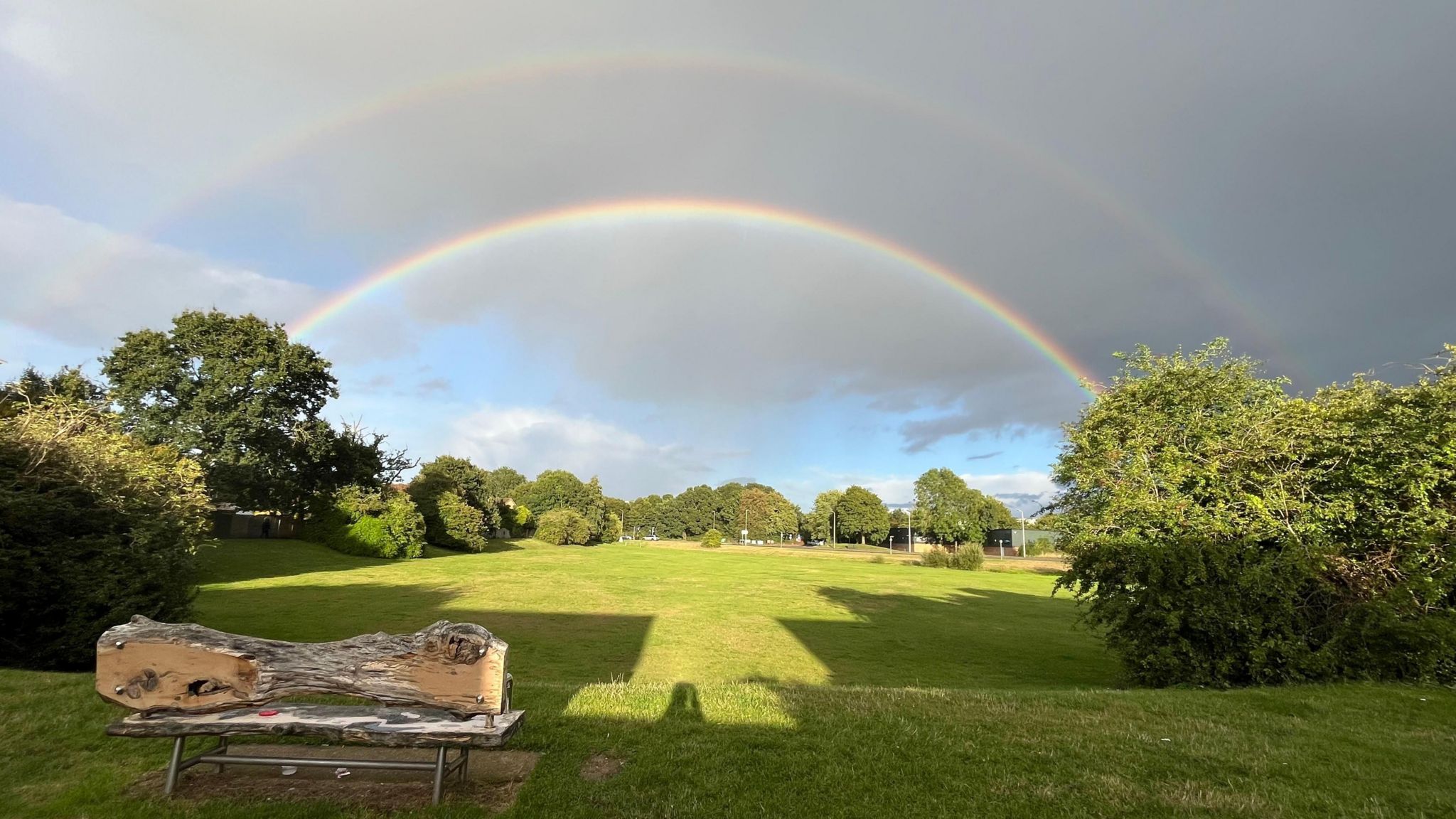 Two rainbows arch over a large green lawn in Thatcham, in front of a mixture of dark grey cloud and blue sky