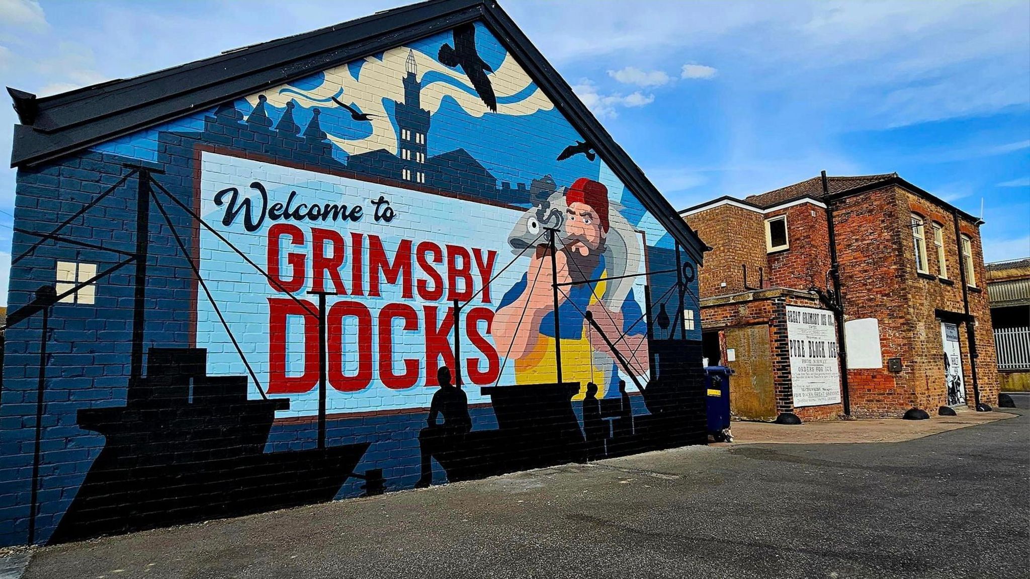 Mural depicting a fisherman with a beard, in yellow overalls and a red hat, holding a fish, there is a blue skyline with boats and bold text that reads: Welcome to Grimsby Docks