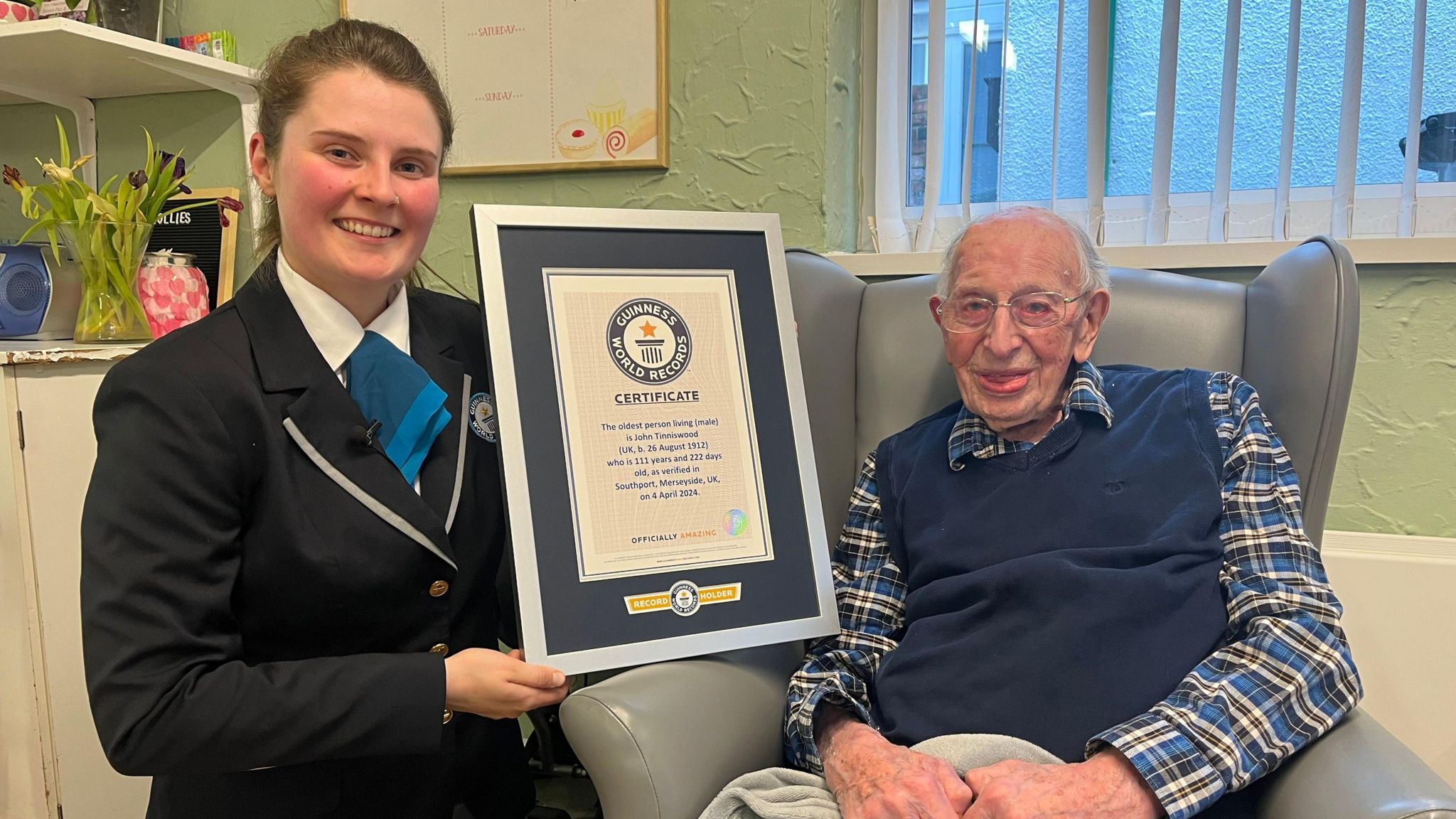 A woman from the Guinness World Records hands a certificate in a frame to John Tinniswood, who is sitting in a grey armchair. Both are smiling at the camera. 