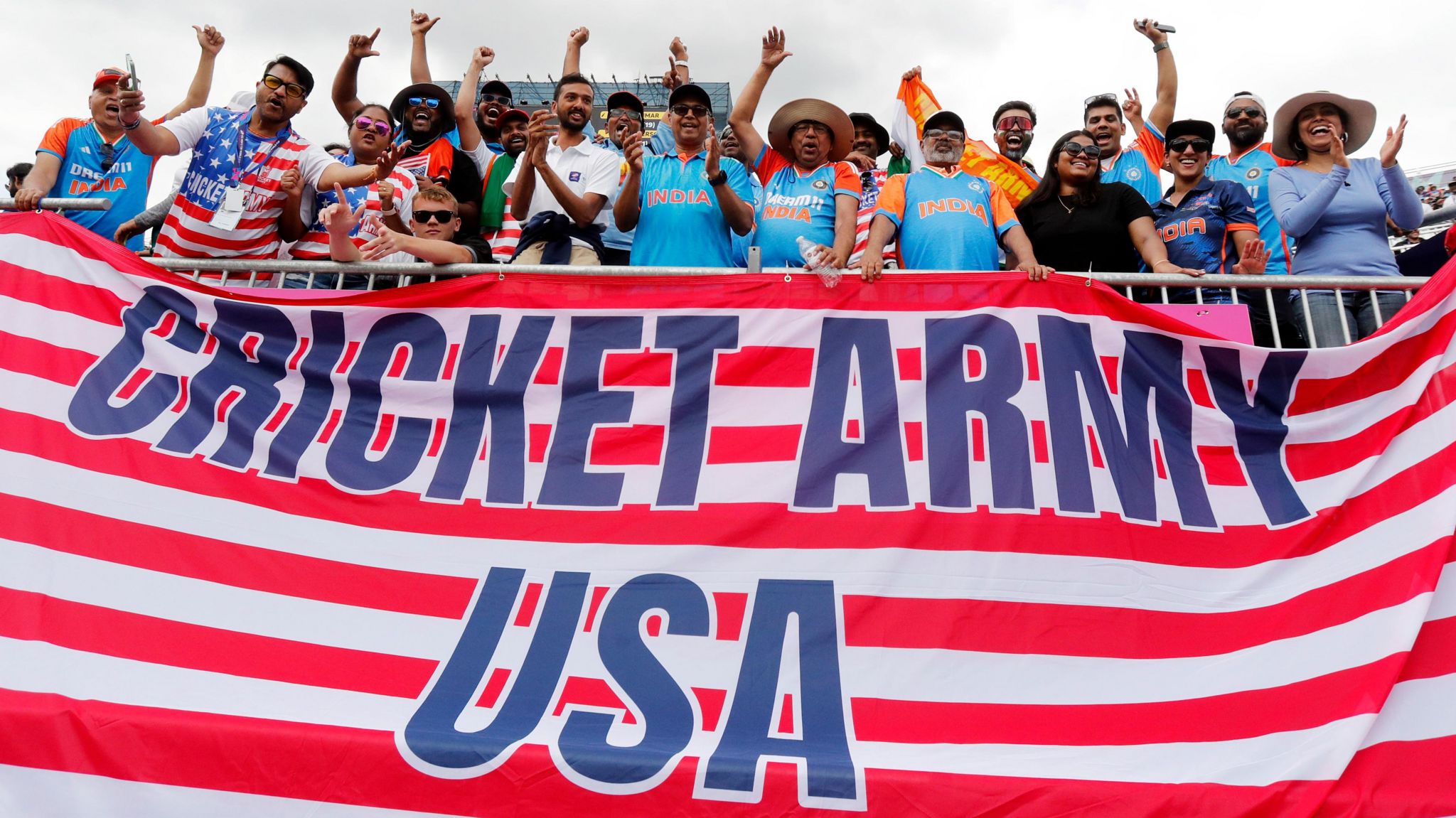 Fans, some wearing India shirts, stand and cheer above an enormous Stars and Stripes flag that has the phrase "Cricket Army USA" written on it