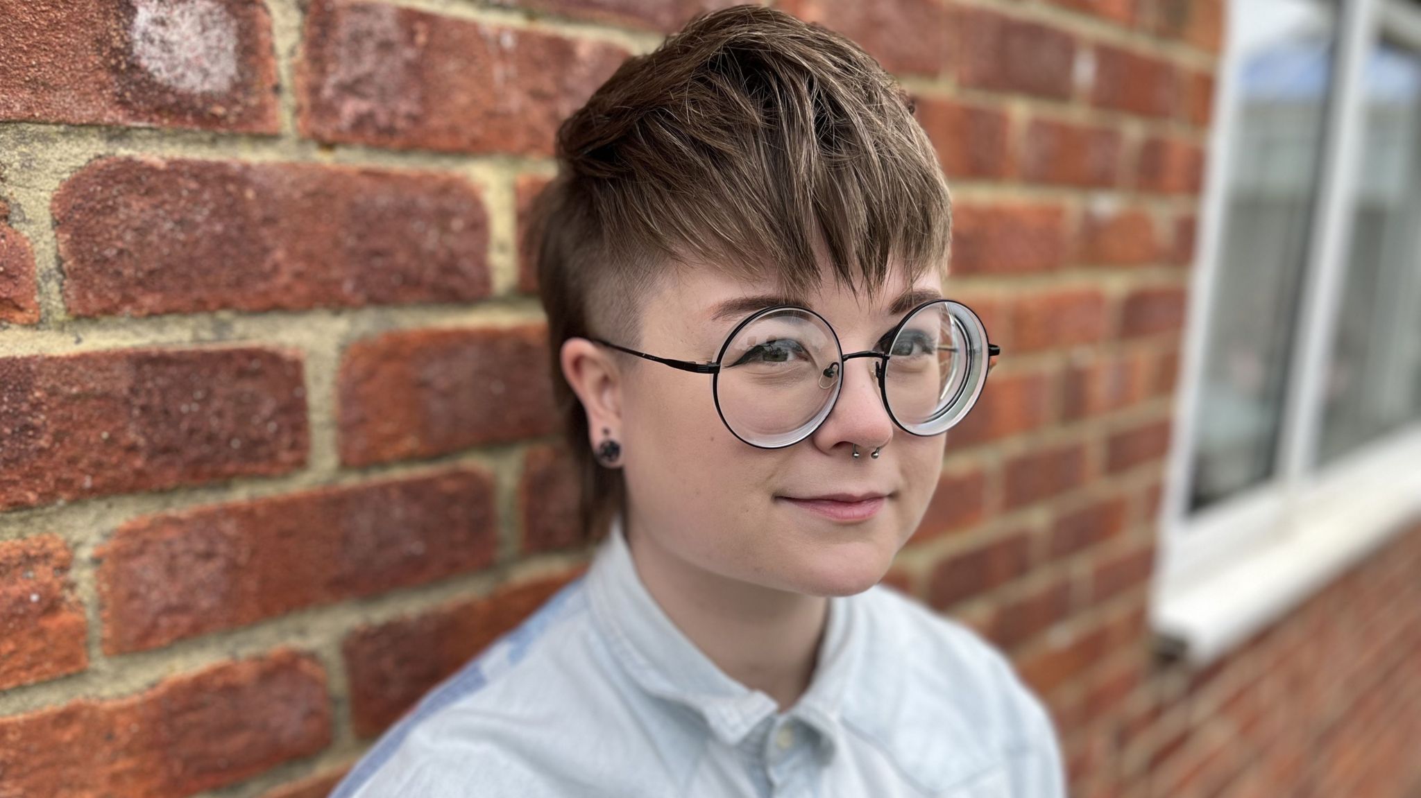 Keesha Brignall standing against a red brick wall wearing round glasses 