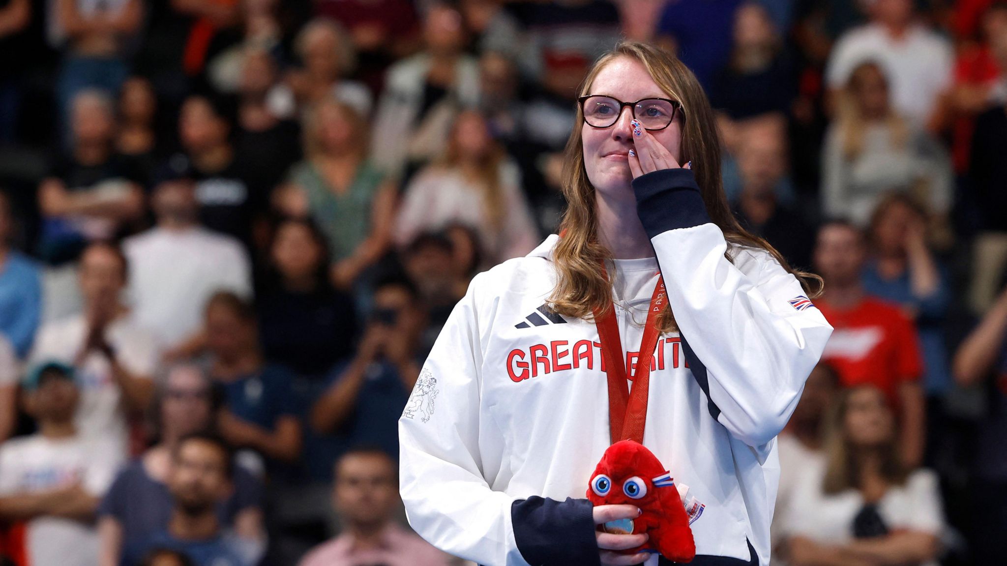 A woman with long brown hair, wearing glasses. She is crying with her hand wiping a tear. 