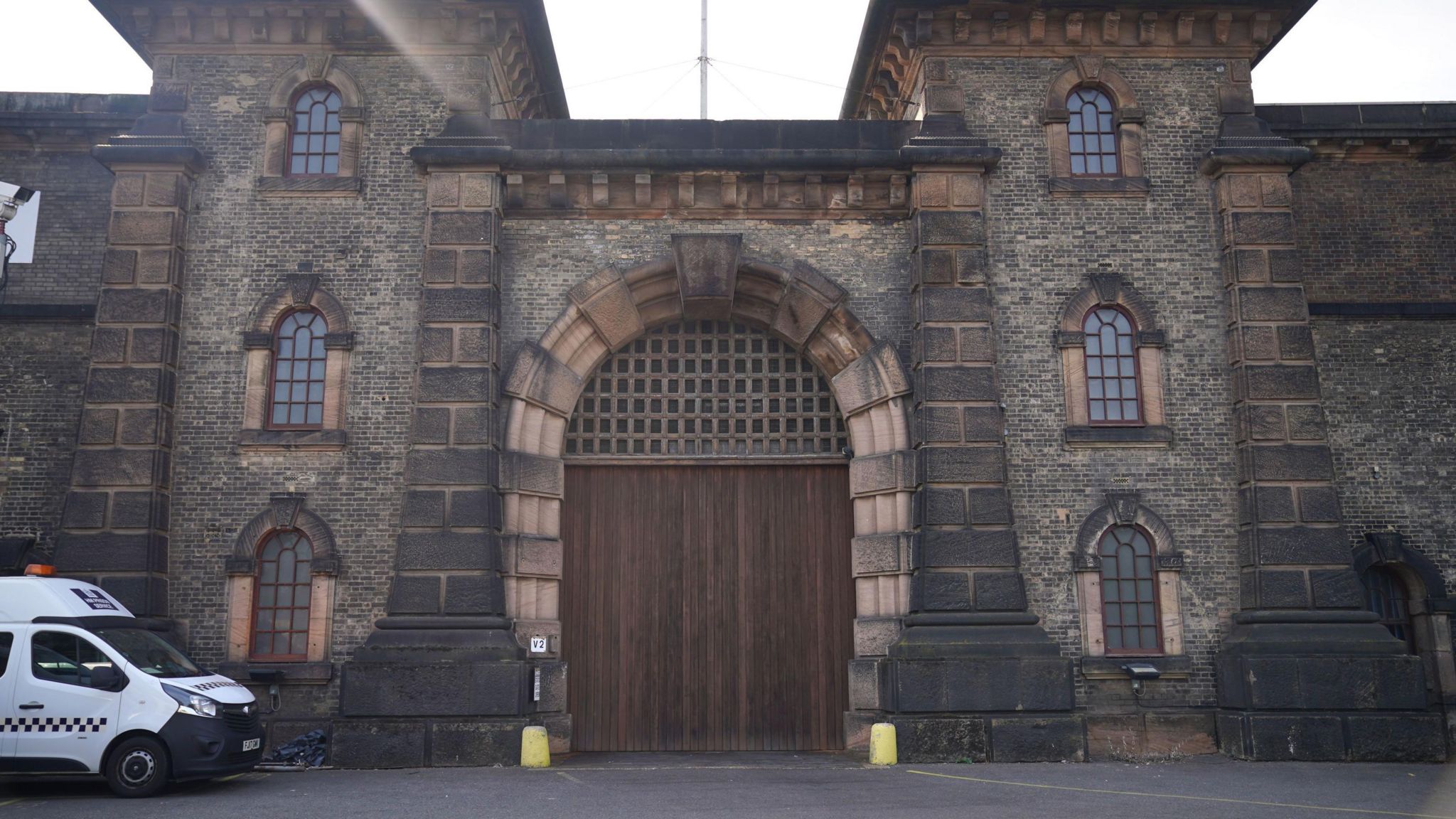 The main entrance to HMP Wandsworth, a Victorian stone building with a large archway entrance with a wooden door and latticed section above. Either side of the door are three arched windows. A police van is parked to the left of the building.