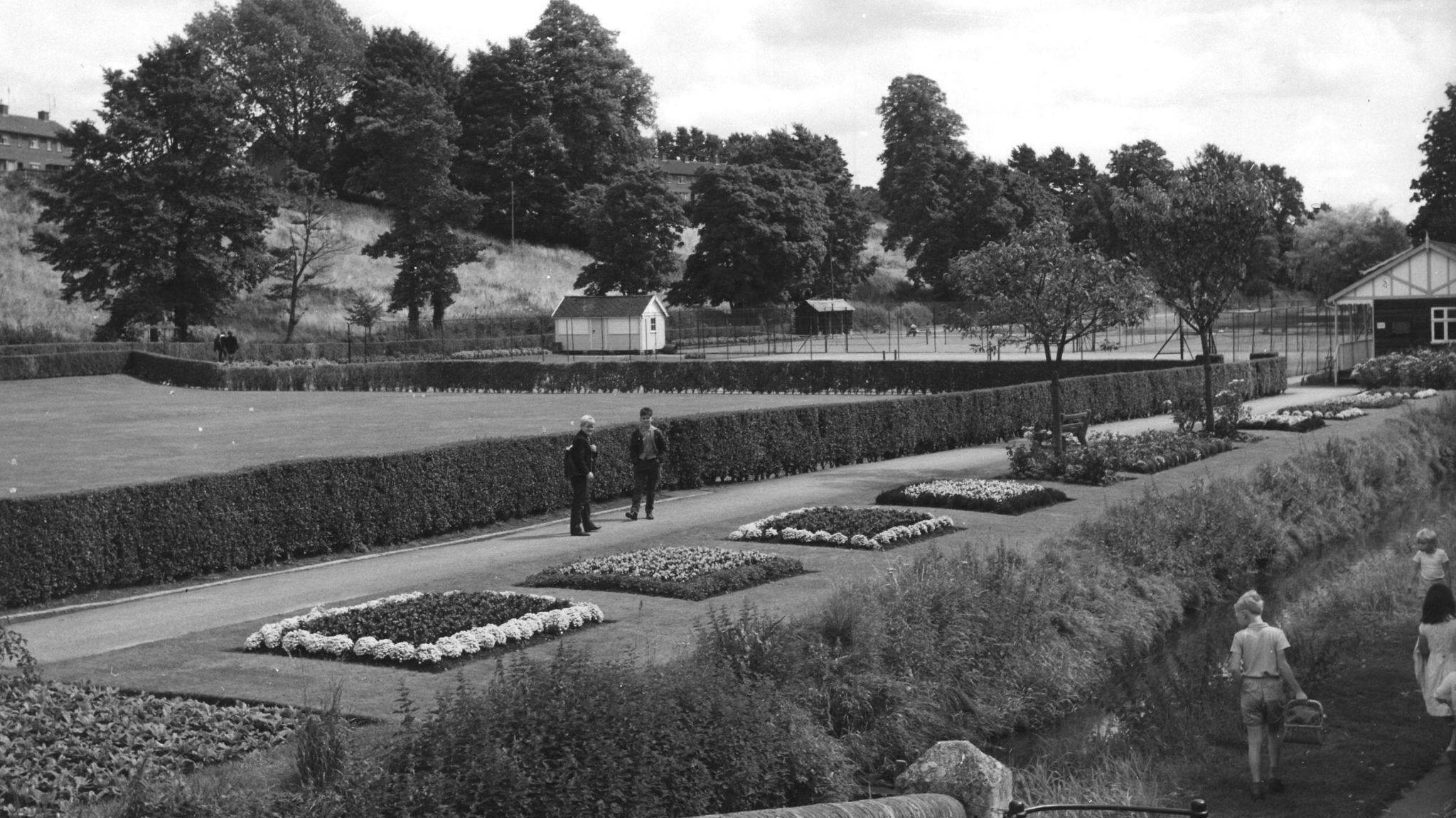 Black and white archive footage of a park. People can be seen walking around it, looking at flower beds. 