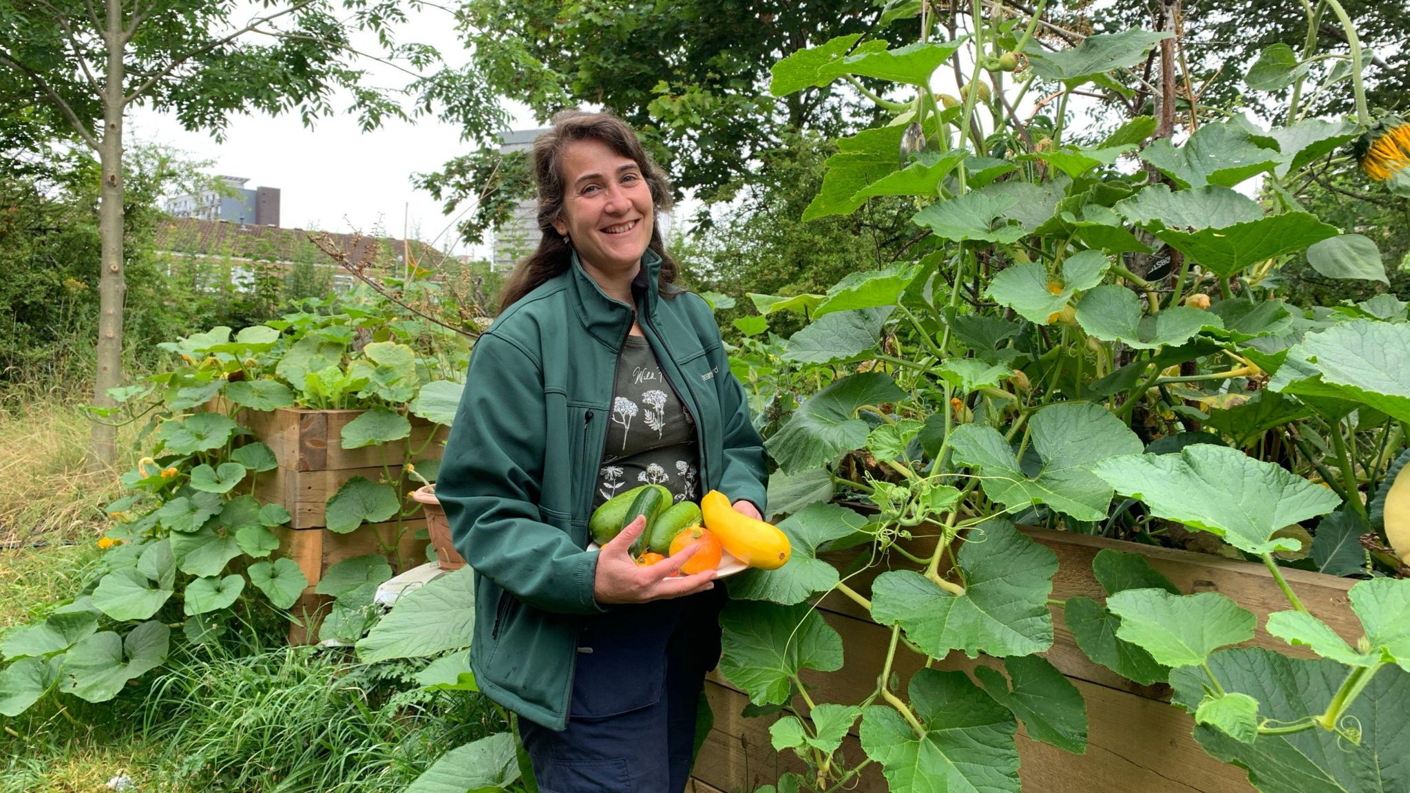 A woman in a dark green jacket holding various gourds whilst stood next to a raised flower bed containing other vegetables