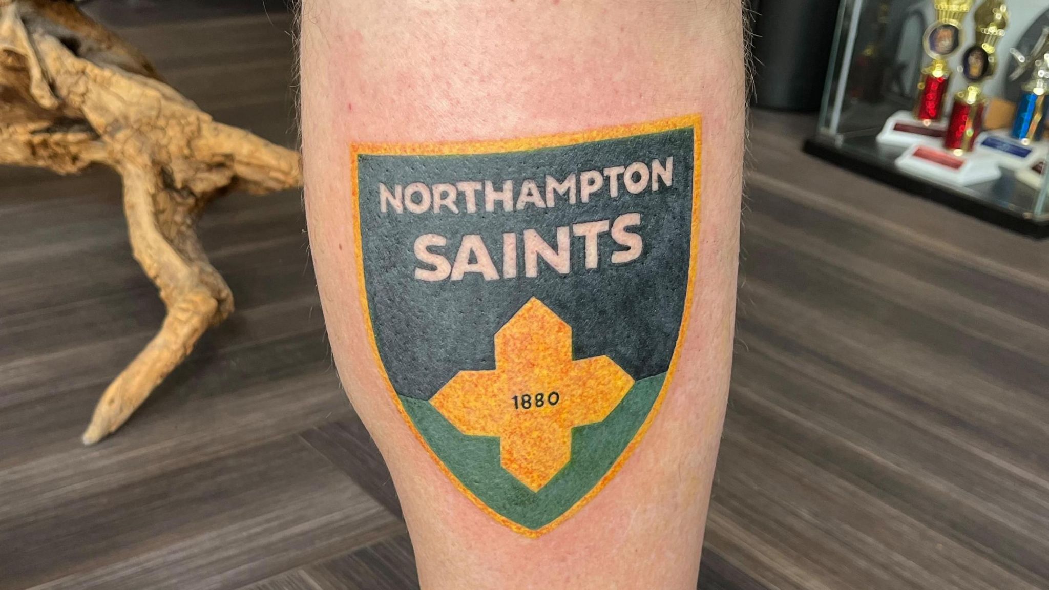 A close-up of a Simon Stacey's leg showing his new tattoo: Northampton Saints rugby club's new crest, which is a shield coloured green, black and gold, a cross, and the words "Northampton Saints".